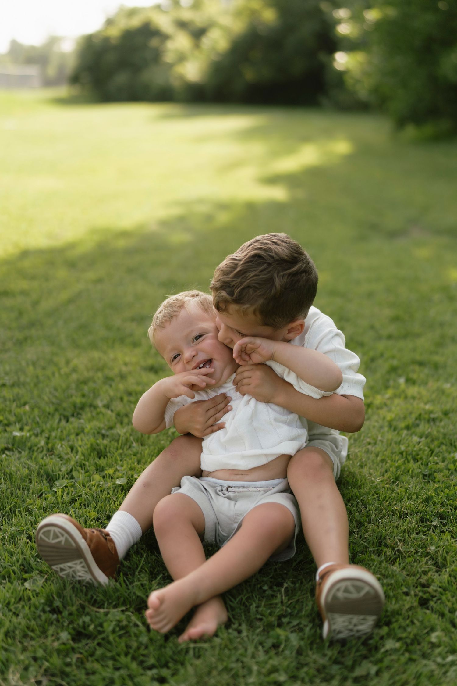 Two young boys sitting on green grass, older boy hugging younger toddler brother outdoors in sunny park.