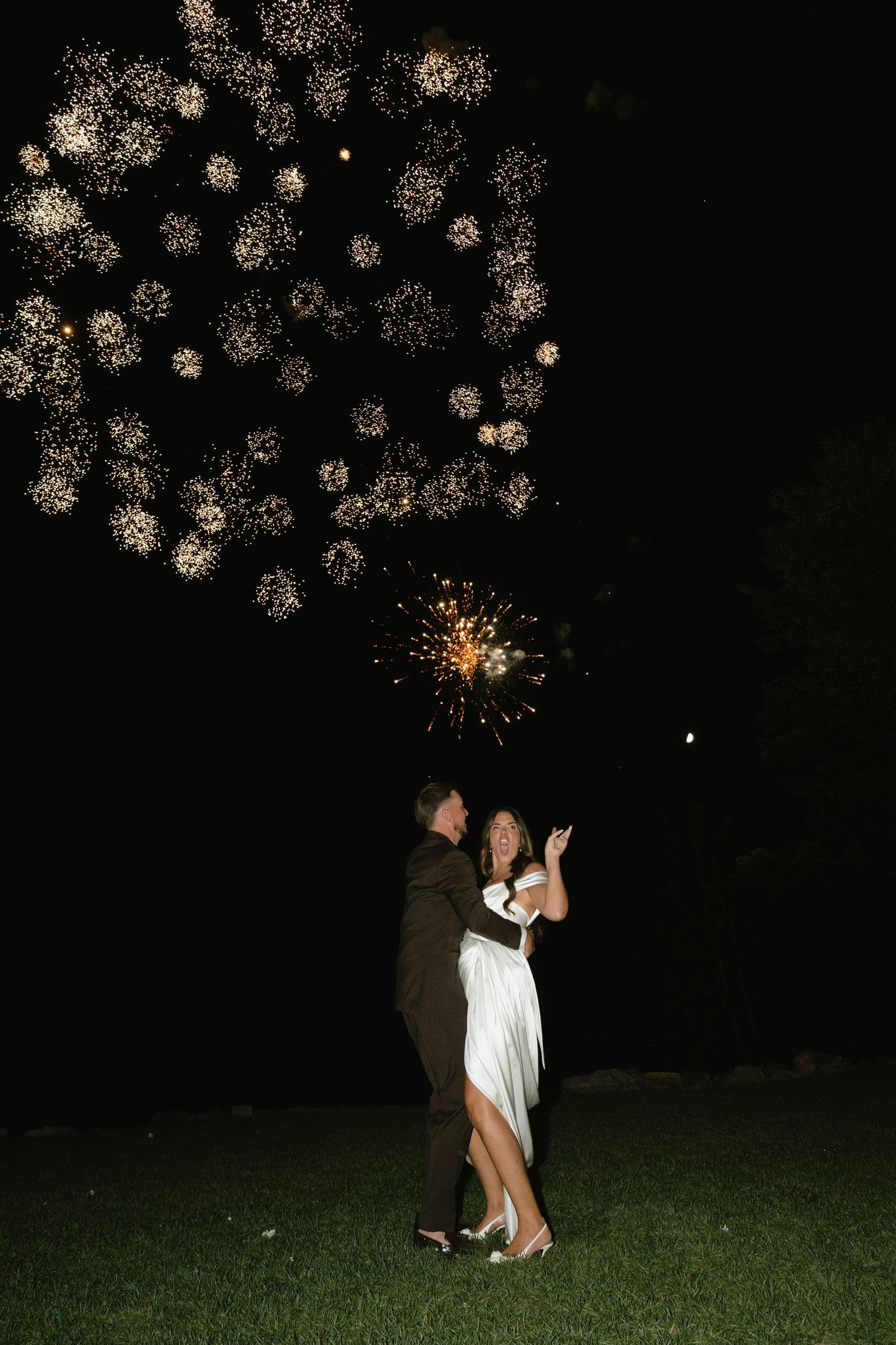 Bride and groom dancing under golden fireworks display at night on green grass at outdoor wedding celebration.