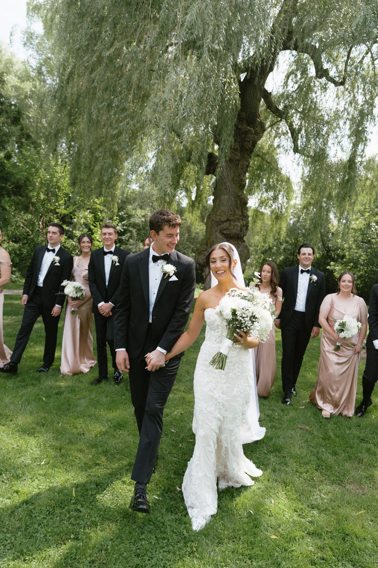 Bride and groom walk together on lush green lawn with wedding party in blush gowns and black tuxedos behind them.
