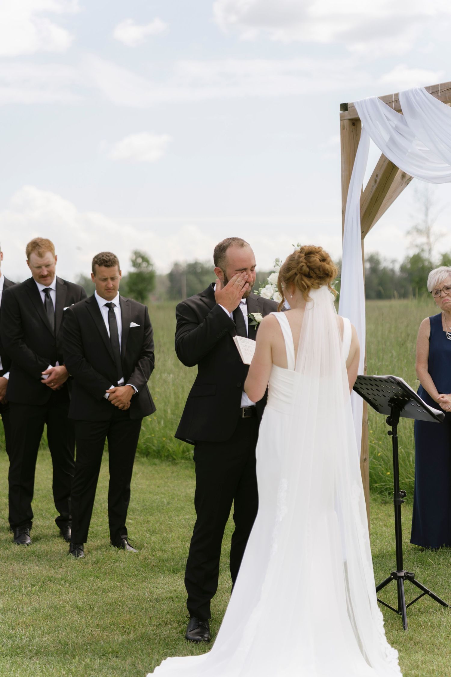 Emotional groom wiping tears as bride stands before him during outdoor wedding ceremony with groomsmen in black suits.