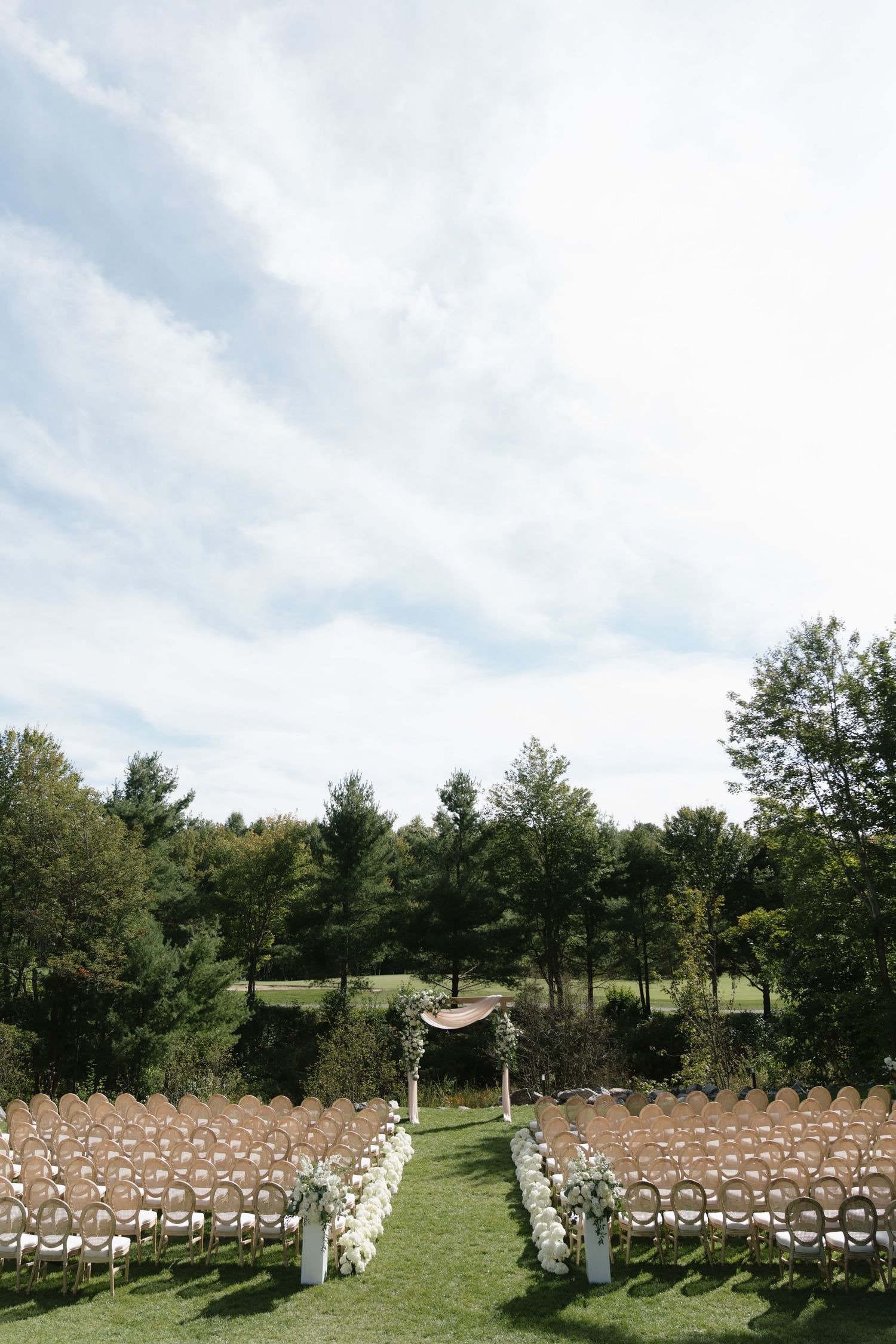 Outdoor wedding ceremony setup with rows of chairs, floral aisle decorations, and an arch amid lush green trees under a blue sky.
