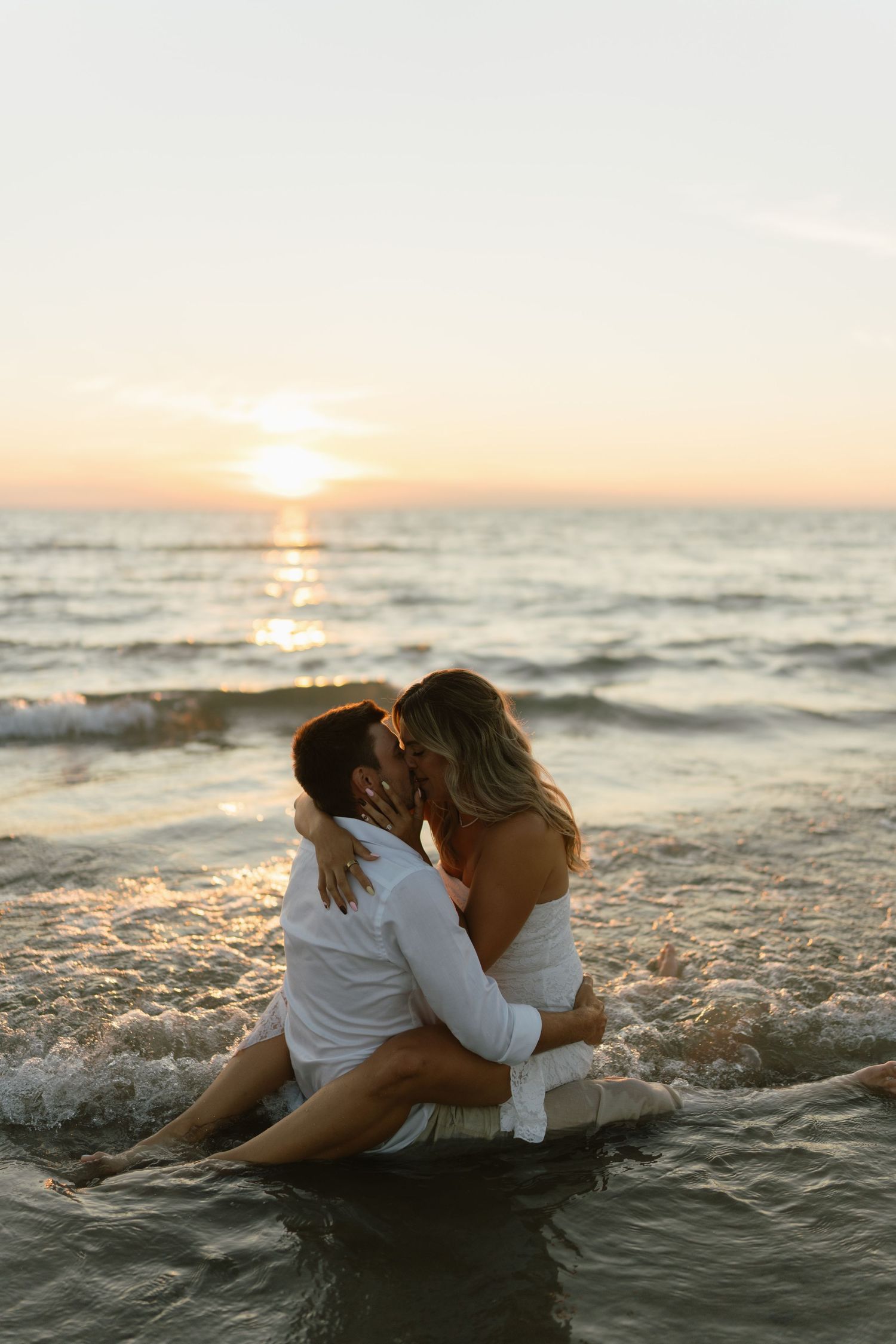 Couple kissing romantically in ocean waves at sunset, sitting in shallow water on a beautiful beach.