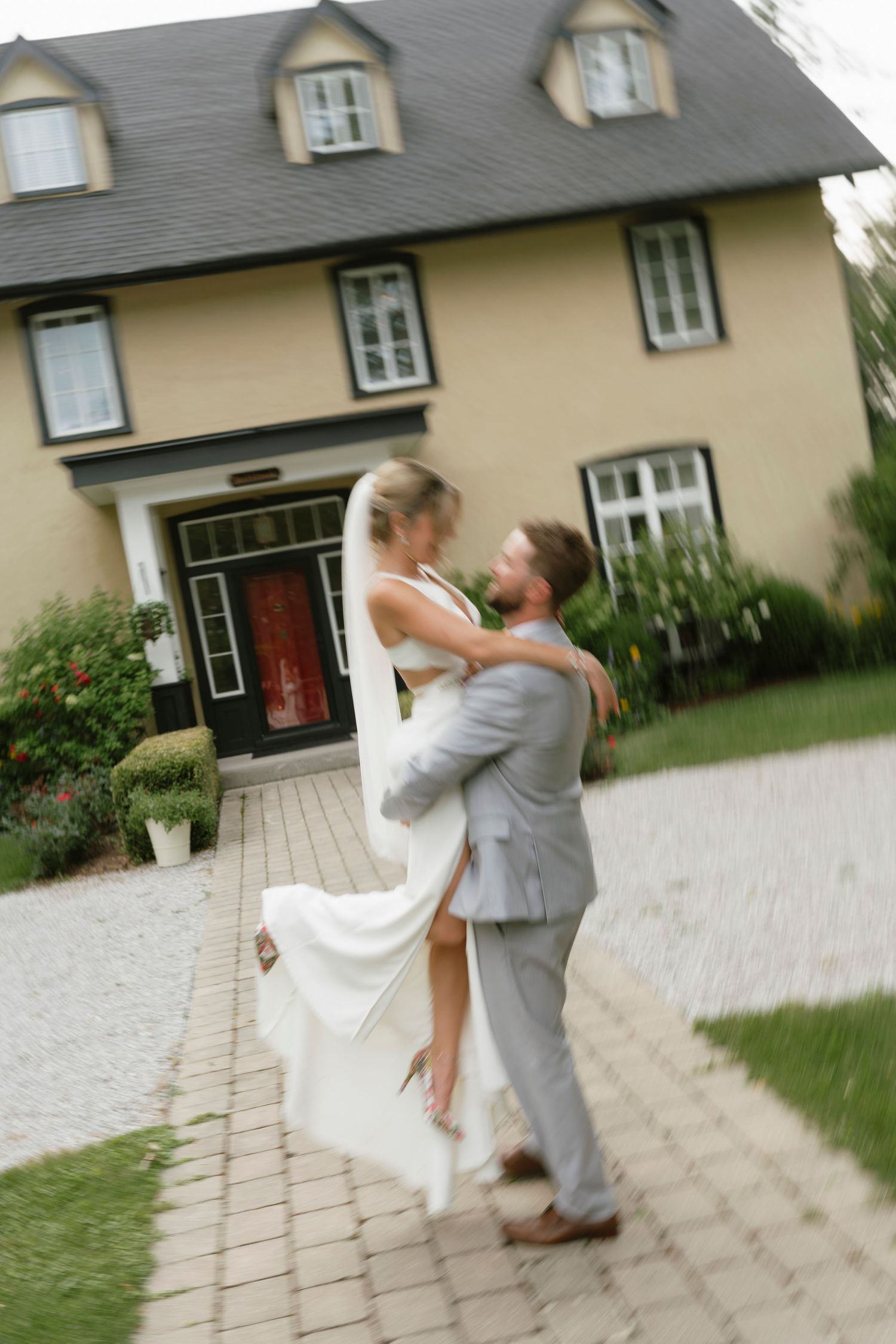 A bride and groom share a romantic embrace on a brick pathway in front of a charming yellow house on their wedding day.
