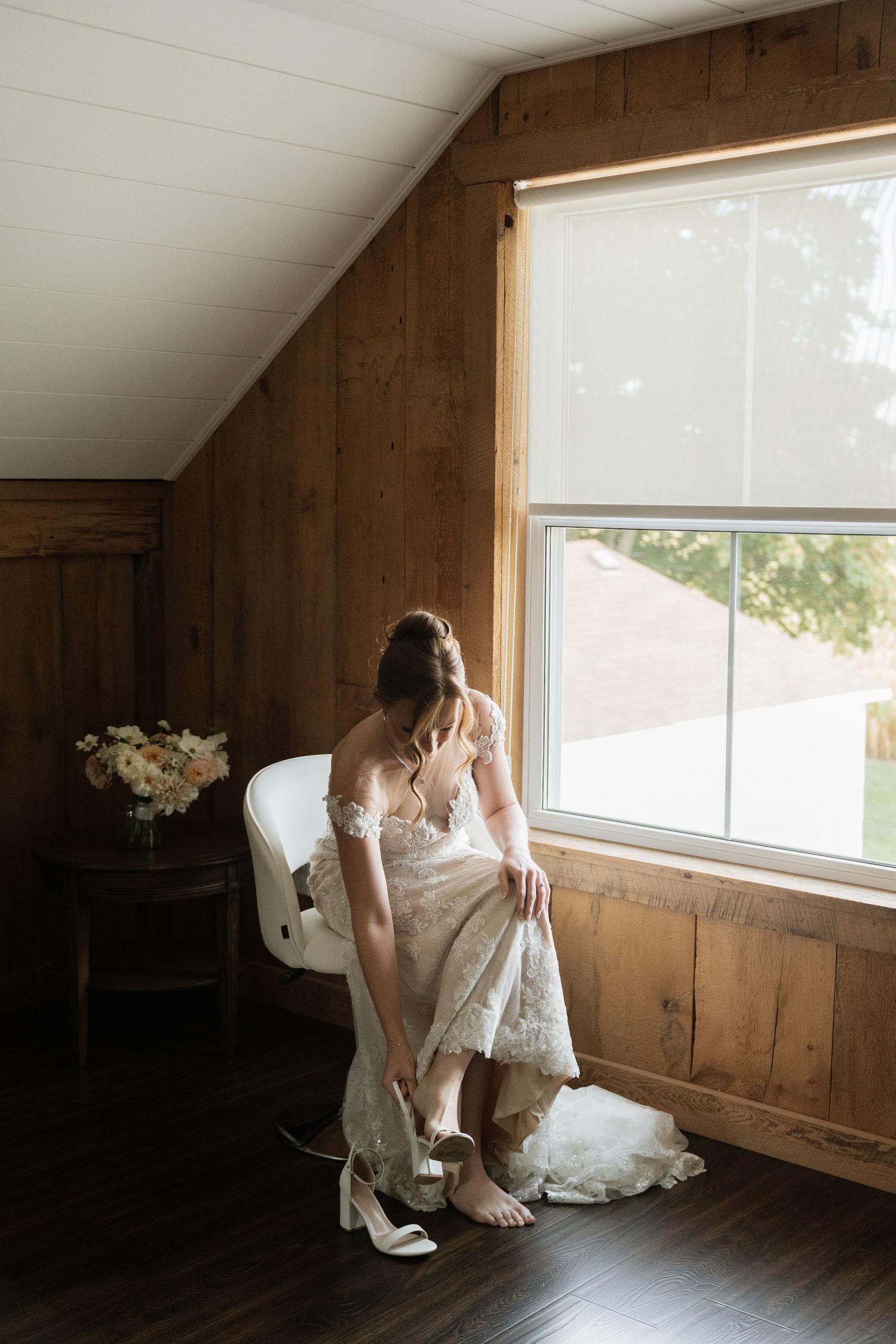A person in a white dress sits alone by a window in a rustic wooden room.