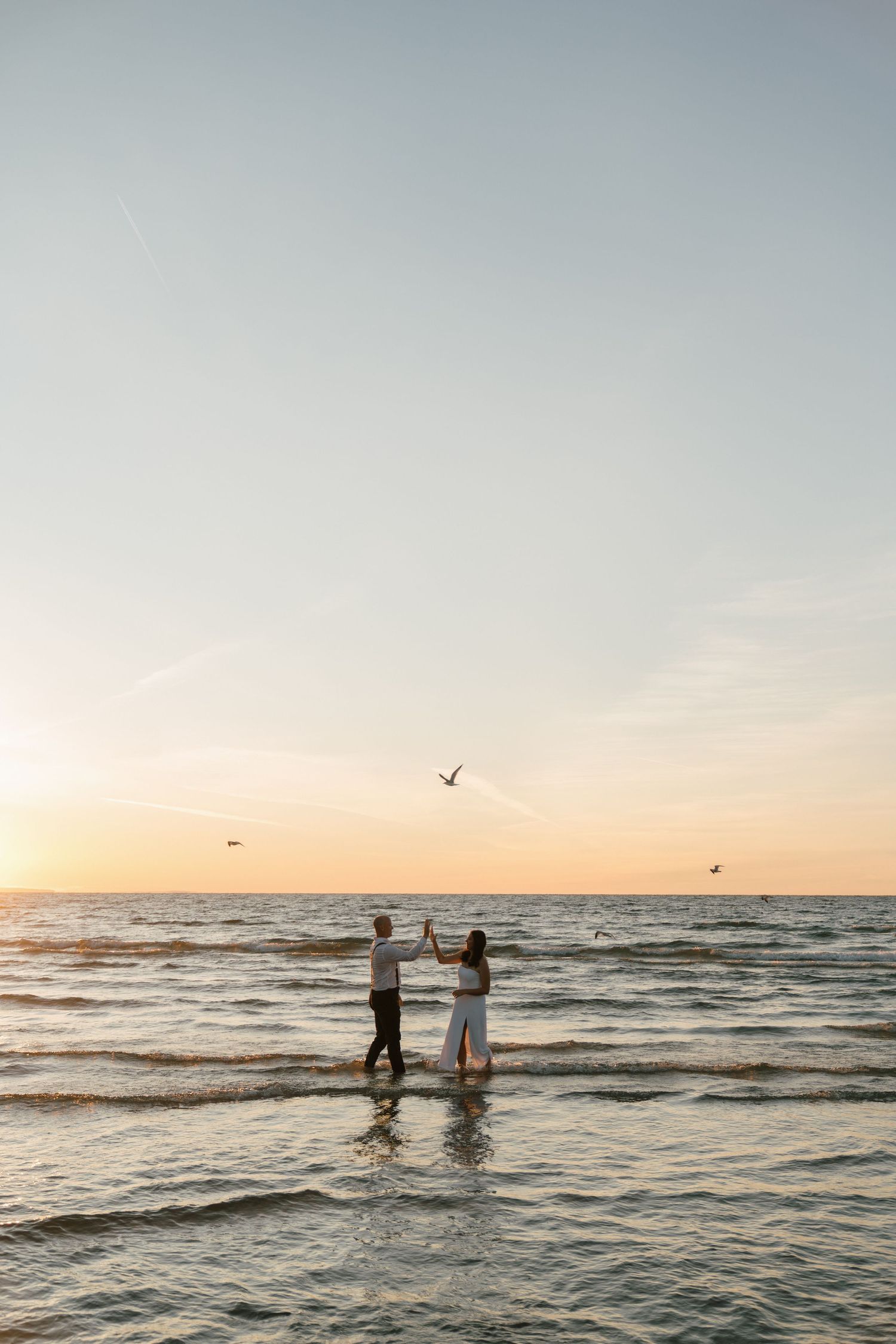 Silhouettes of people wade in ocean waves at sunset as seabirds fly overhead.