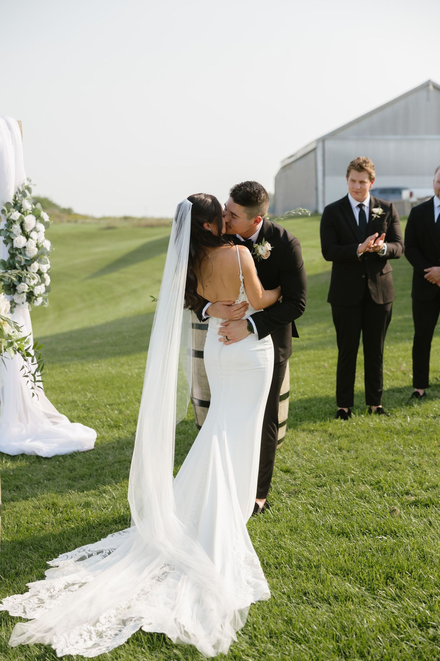 Bride and groom share first kiss at outdoor wedding ceremony on lush green lawn with floral arch.