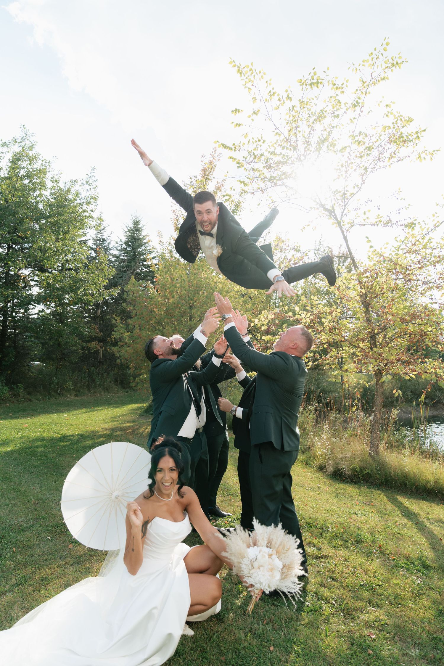 Groomsmen toss groom in air while bride watches during outdoor wedding photos.