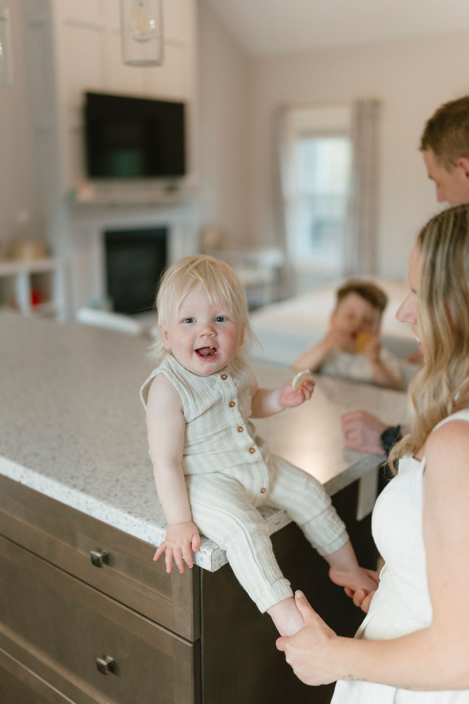 A smiling blonde toddler sits on a kitchen counter in a striped romper while family members gather in a bright modern home.