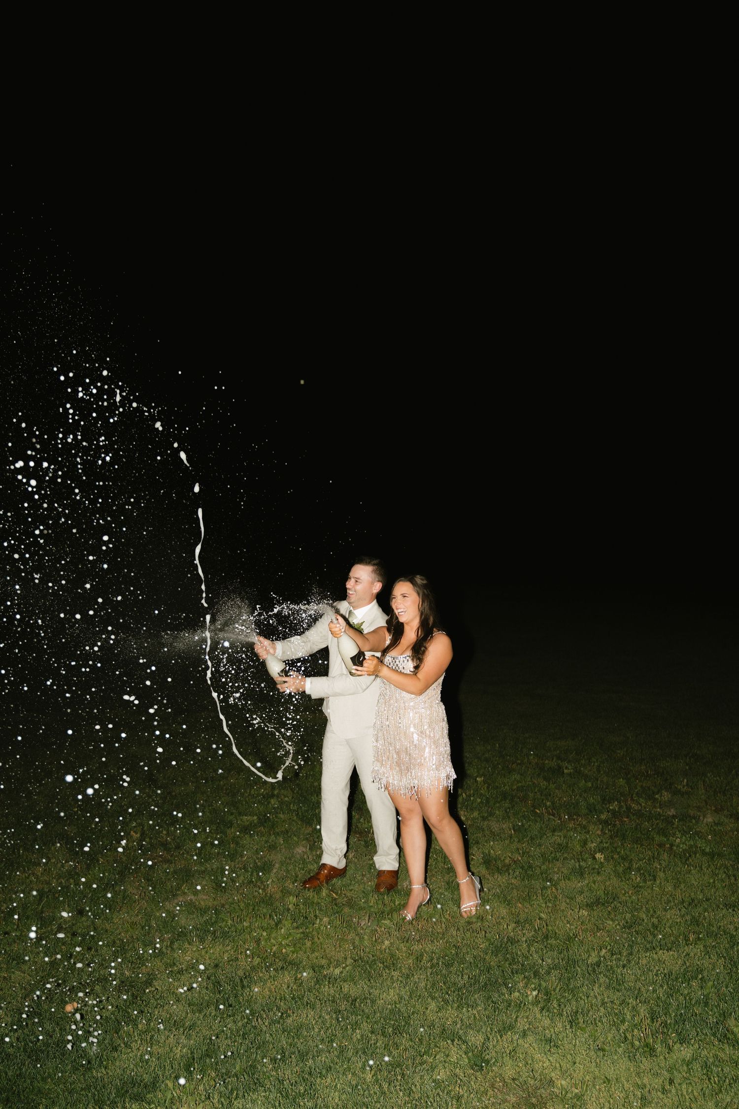 People celebrate by spraying champagne in the dark on a grassy field.