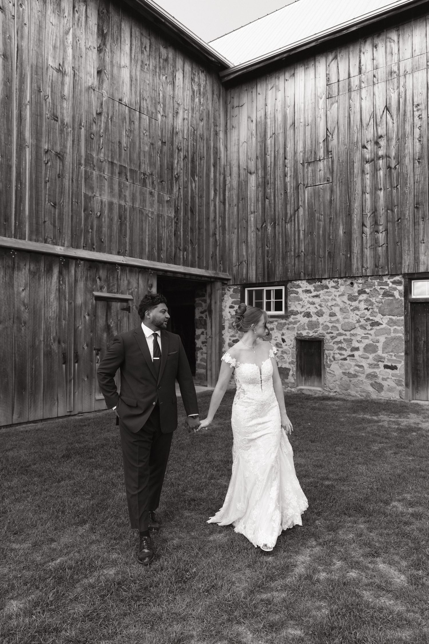 A couple walks together outside a rustic barn venue in black and white.