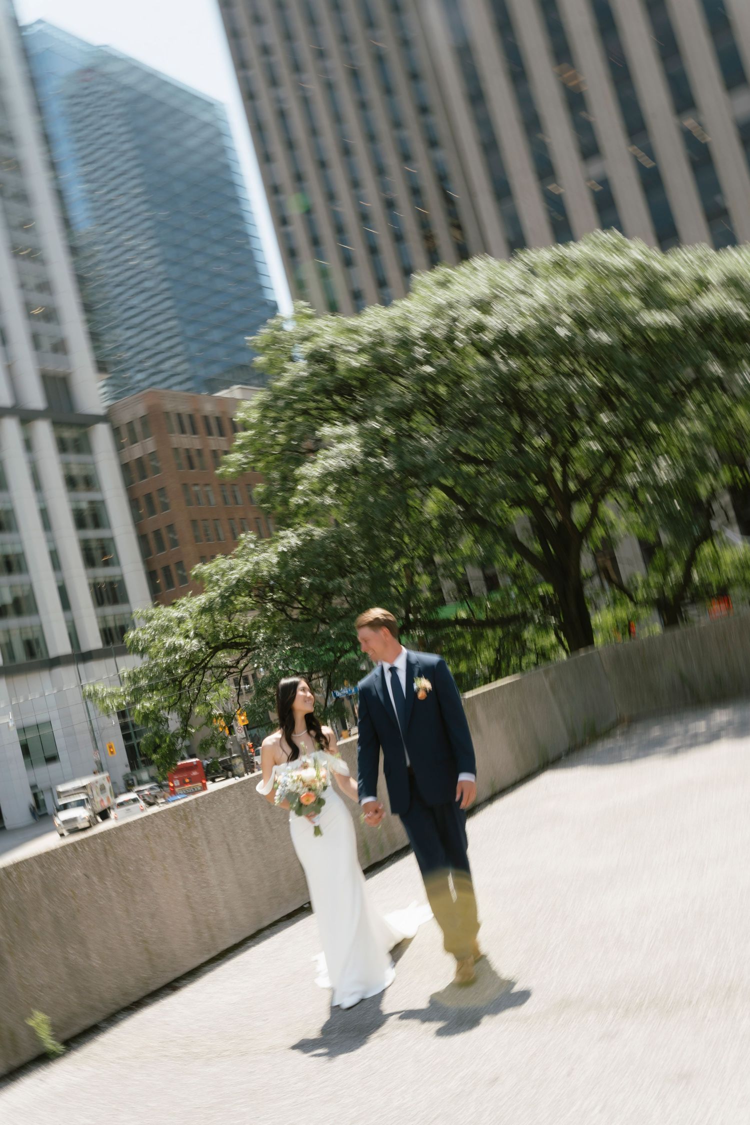 Bride and groom walk hand-in-hand through a sunny urban plaza surrounded by skyscrapers and green trees.