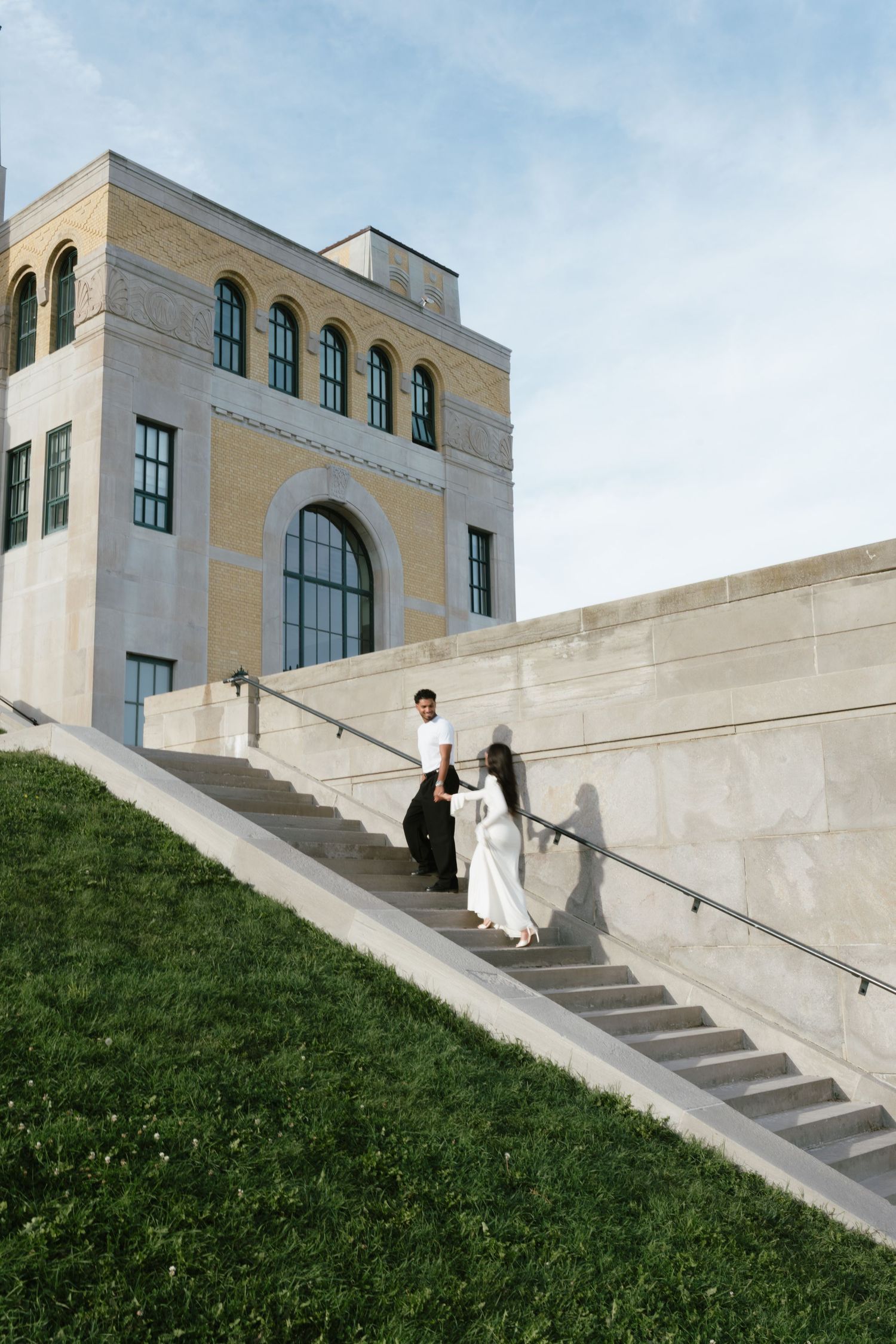 Couple in wedding attire descending outdoor stone stairs beside a classic brick building with arched windows.