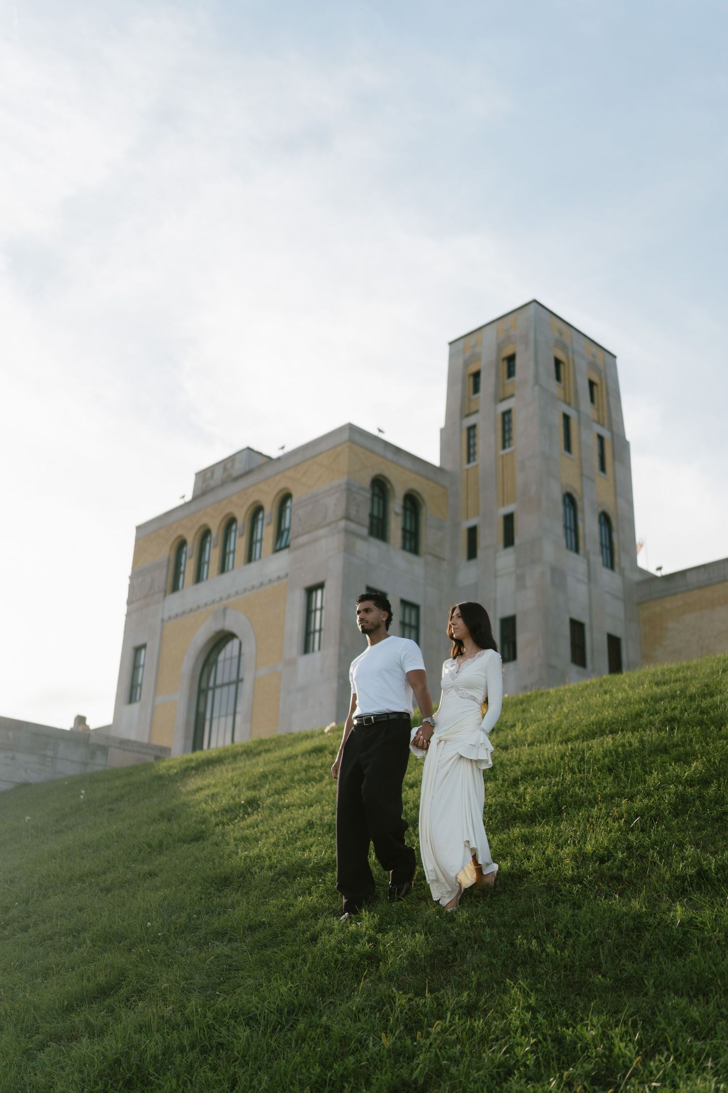 Couple holding hands on a grassy hill in front of a historic stone building under a cloudy sky.