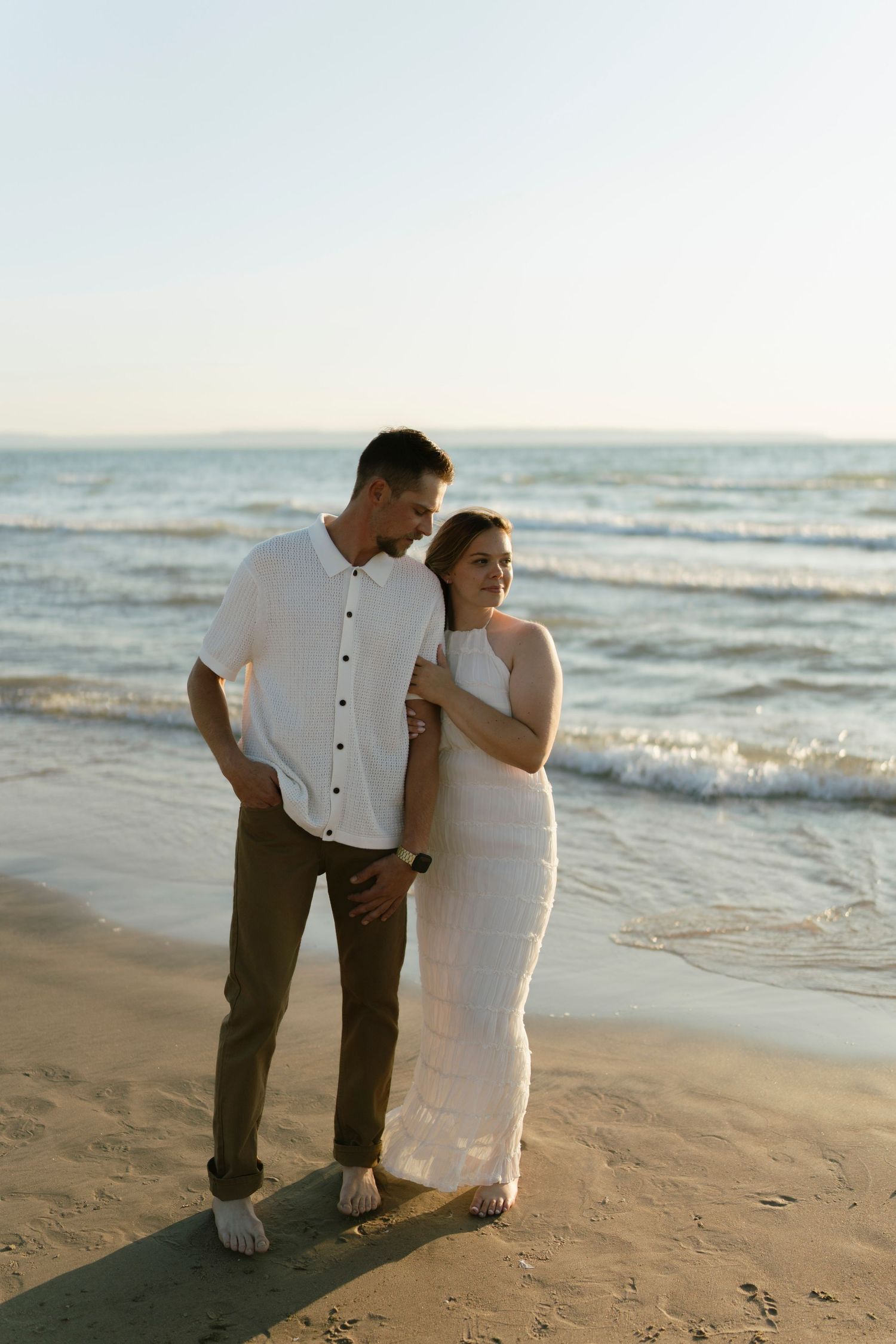 Couple in white outfits standing barefoot on a sandy beach at sunset, with ocean waves in the background.