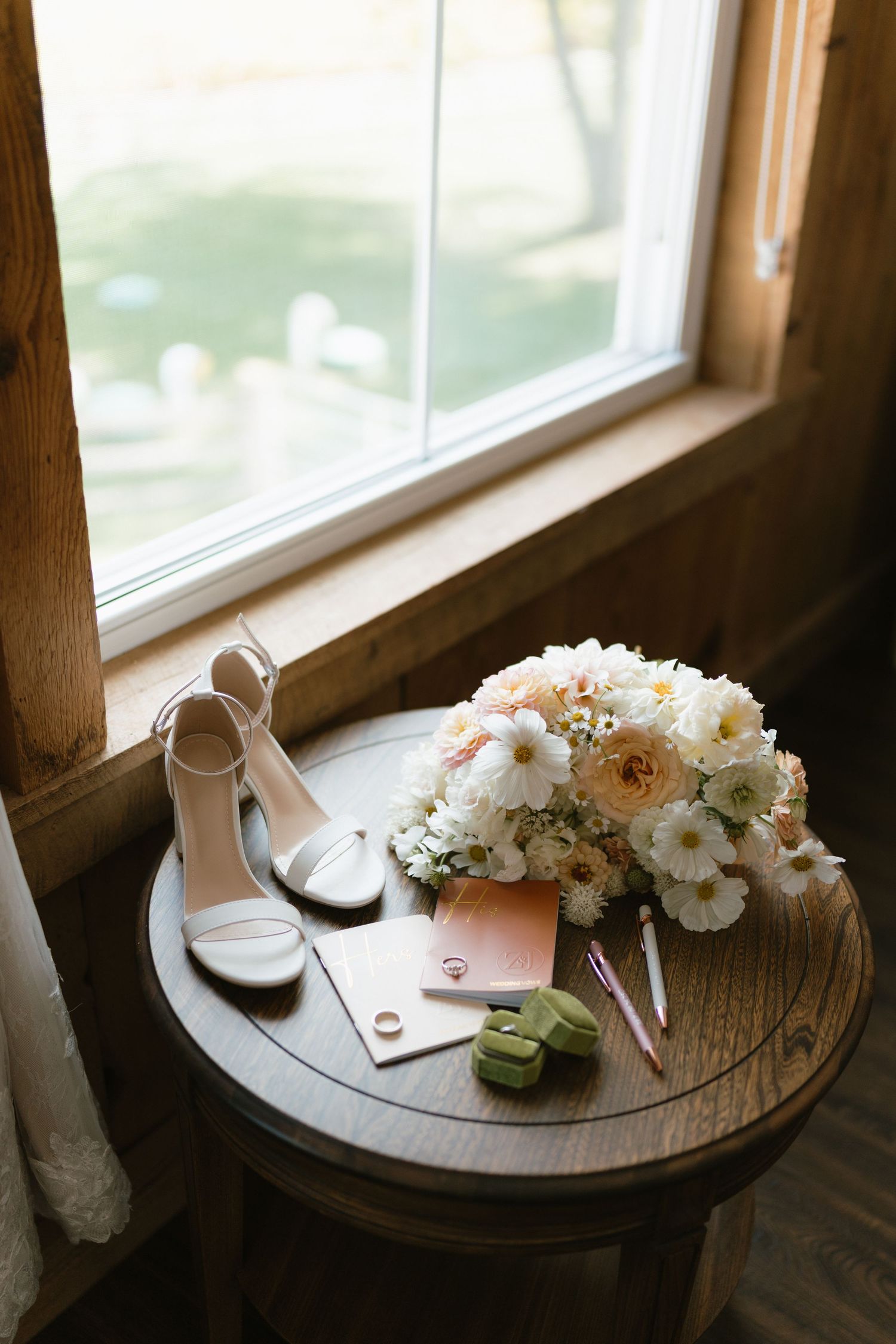 Wedding details including bridal shoes and floral bouquet arranged on wooden table by window.
