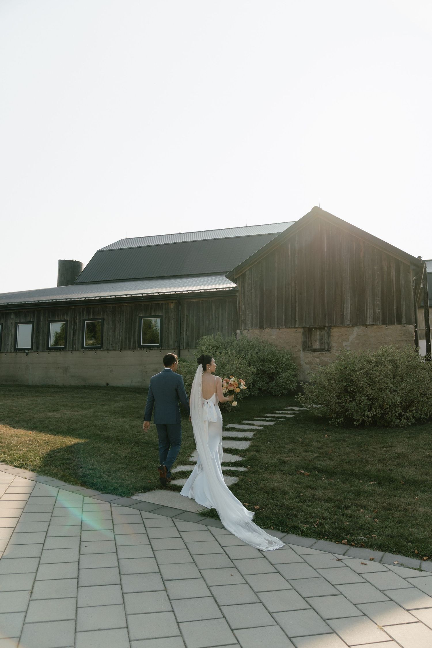 A couple walks along a stone pathway outside a rustic barn venue at sunset.
