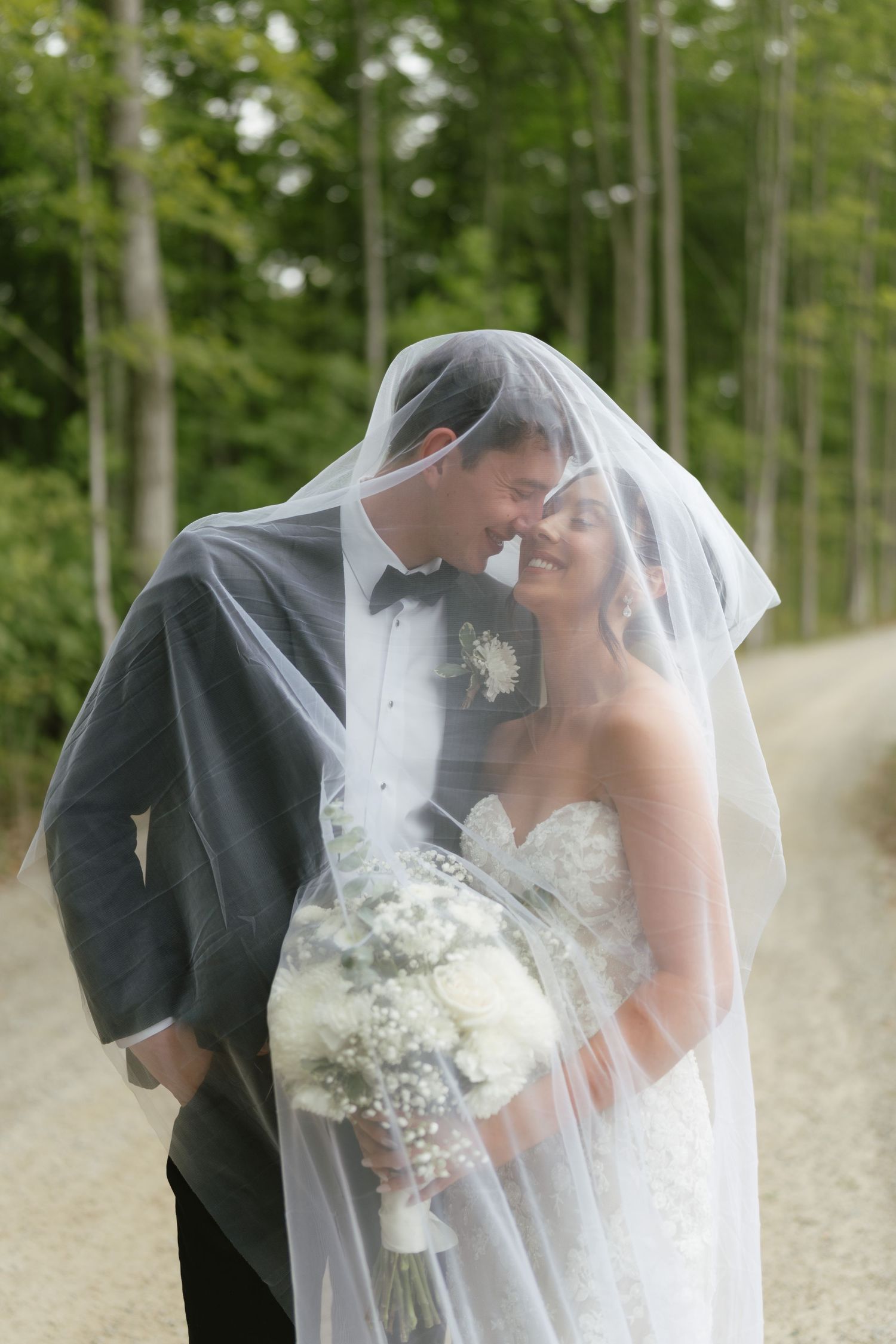 Bride and groom share romantic moment under wedding veil on tree-lined path, bride holding white bouquet.