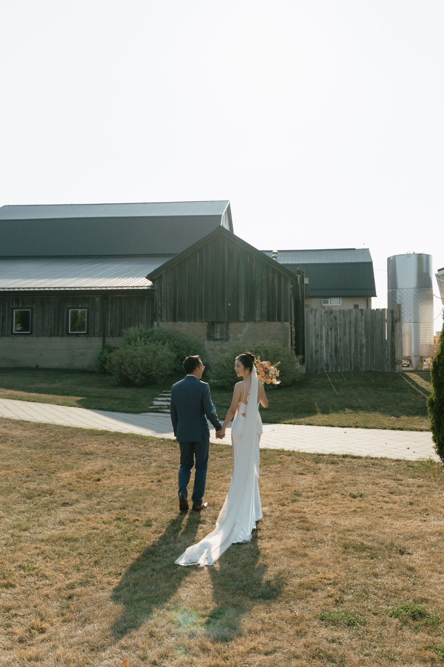 Newlyweds walking hand in hand near a rustic barn on their wedding day.