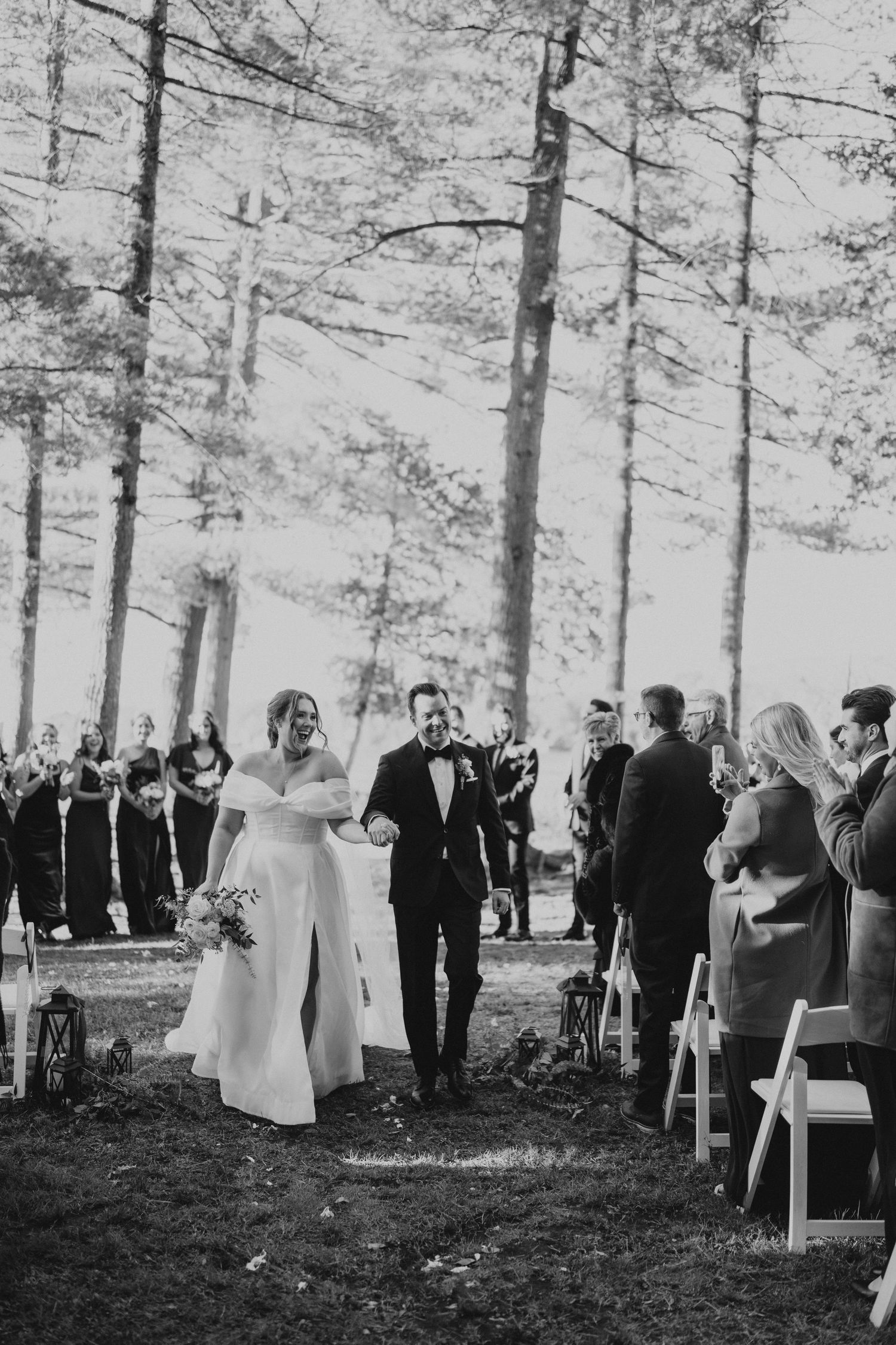 Outdoor forest wedding ceremony with guests seated on white chairs among tall pine trees.