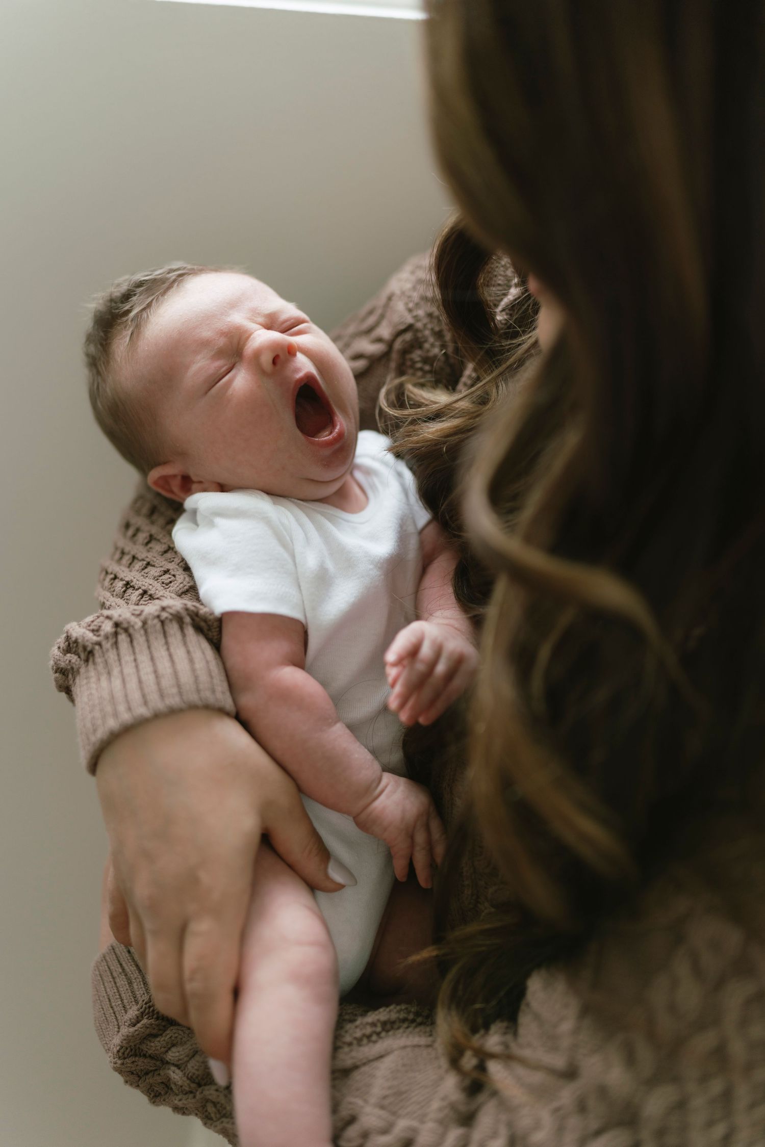 A mother in a knit sweater cradles her yawning newborn baby dressed in a white onesie.