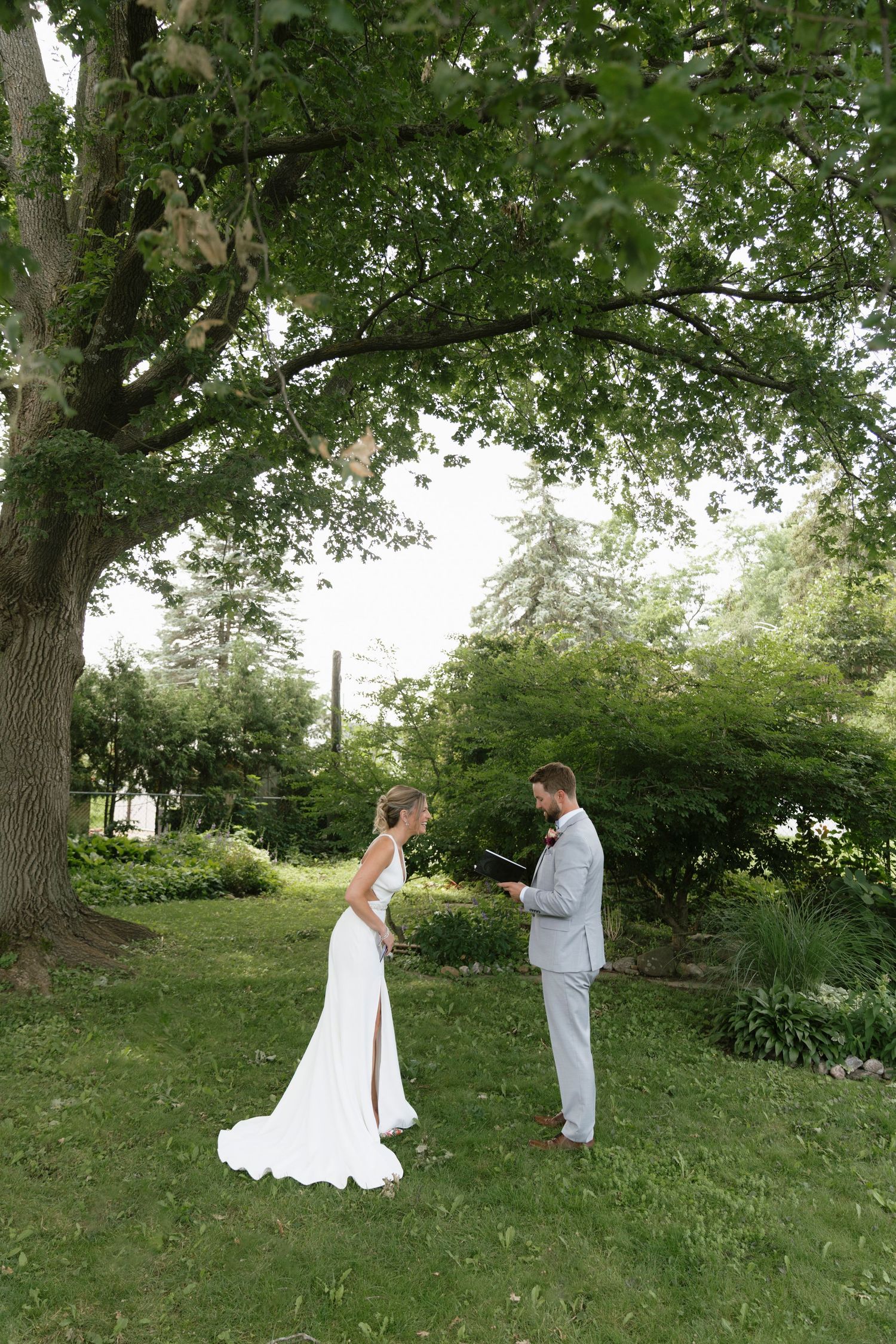A bride and groom exchange vows outdoors under a large green tree in a lush garden setting.