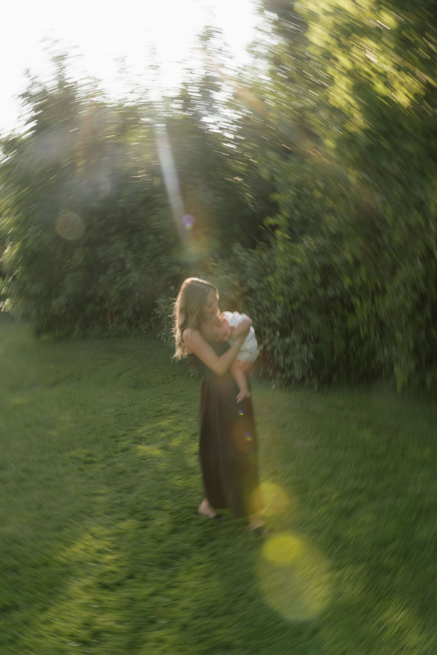 Mother holding baby in sunlit green garden, wearing dark dress, with dreamy bokeh lens flare effect.