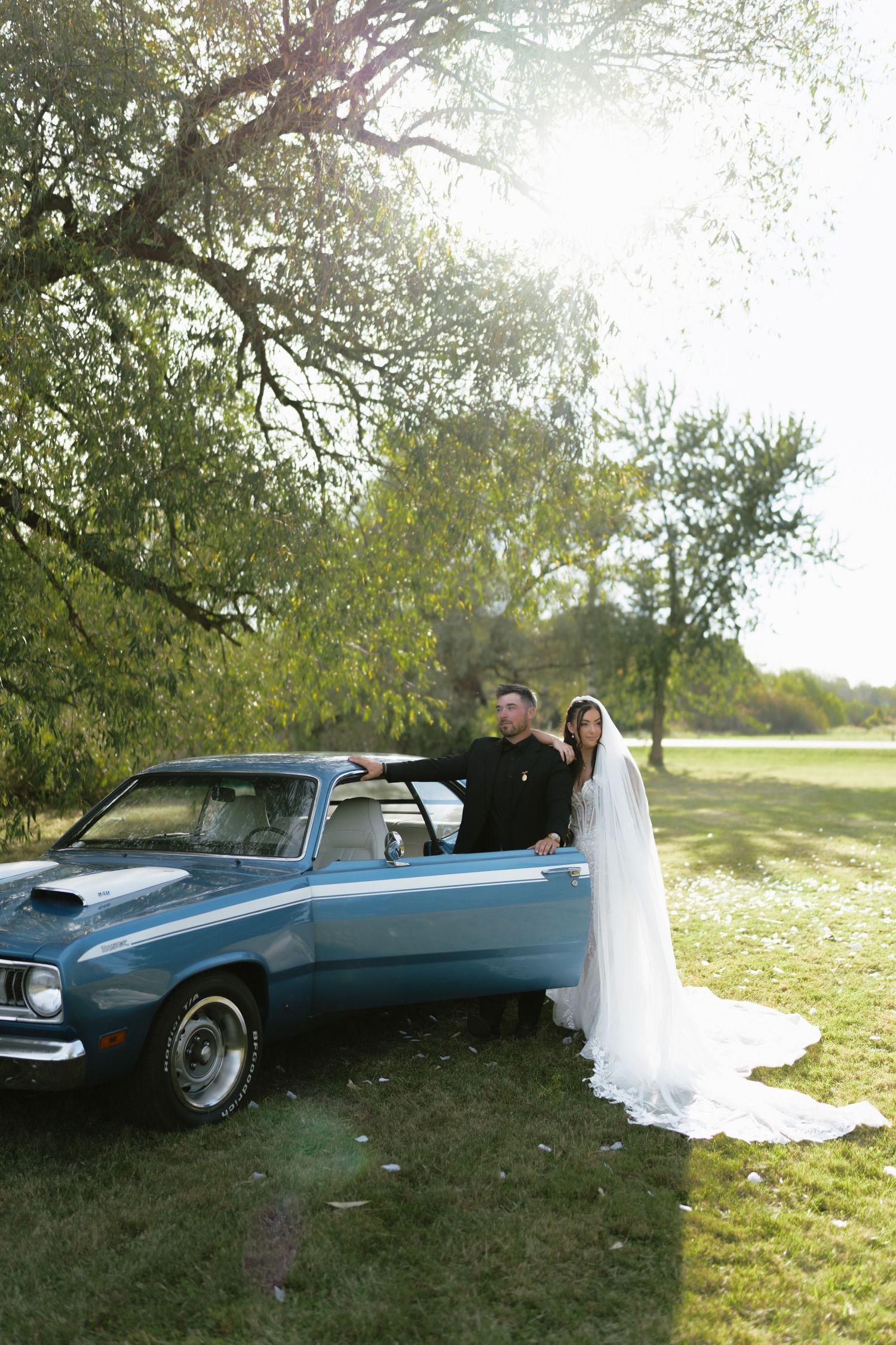 Bride in white veil and groom pose beside a classic blue convertible car under a sunlit tree outdoors.
