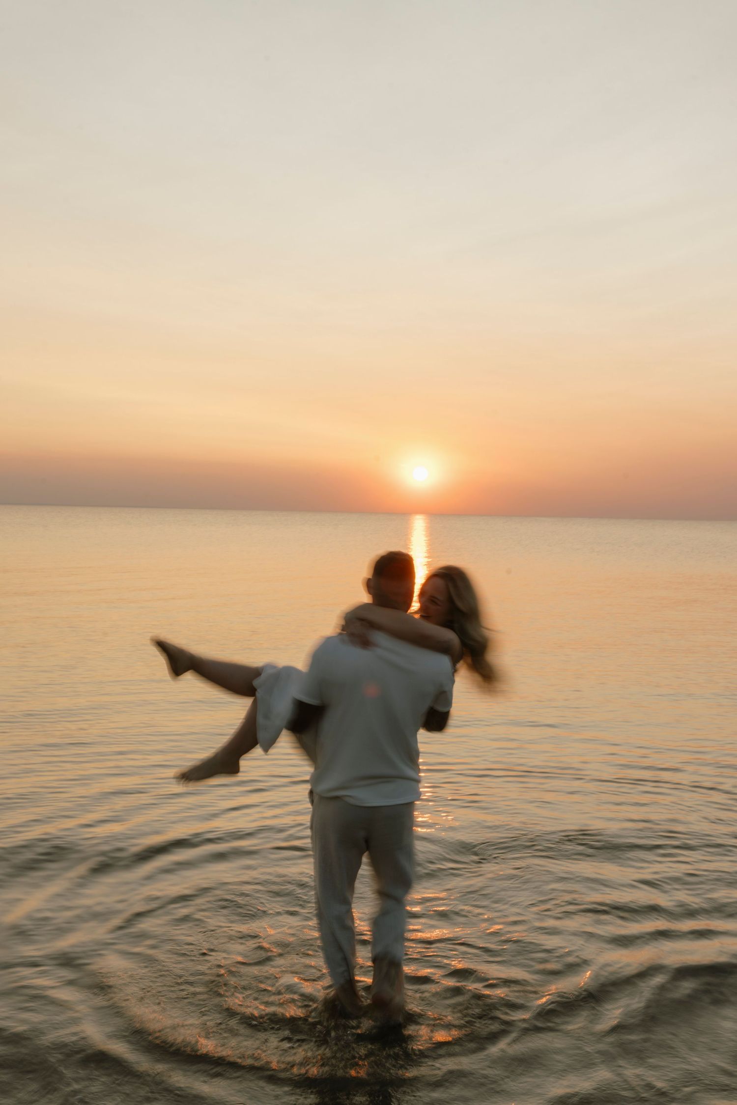 A couple embraces at sunset, standing in shallow ocean water, with a warm golden sky reflecting on the sea.
