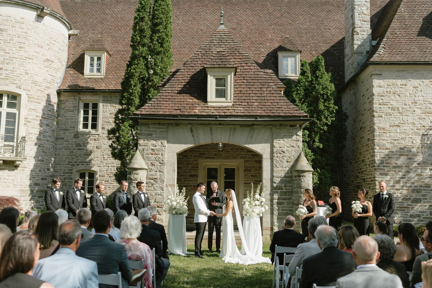 Outdoor wedding ceremony at stone castle with guests seated on lawn.