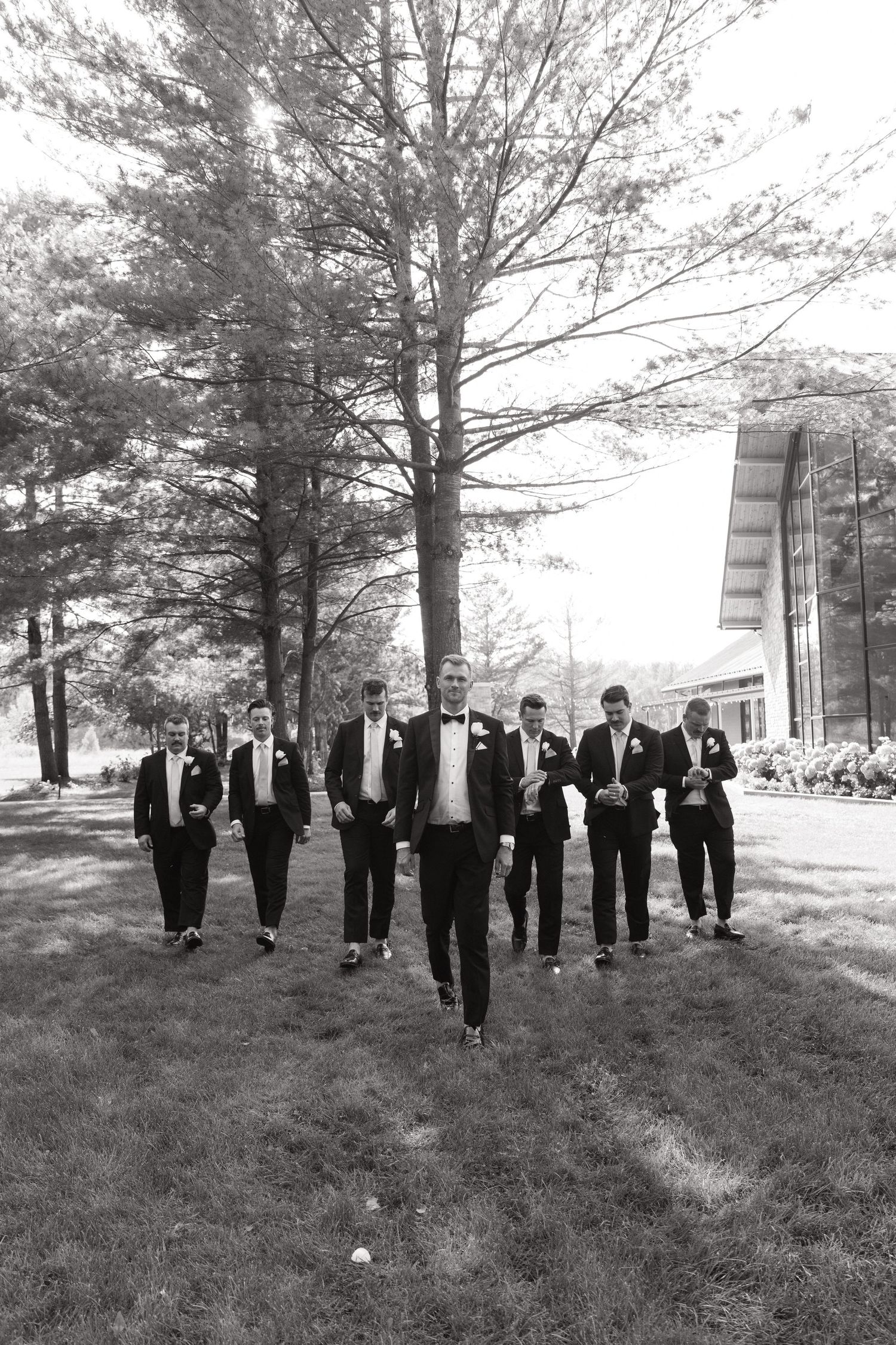 A group in formal black tuxedos walks across a grassy lawn in black and white photography.