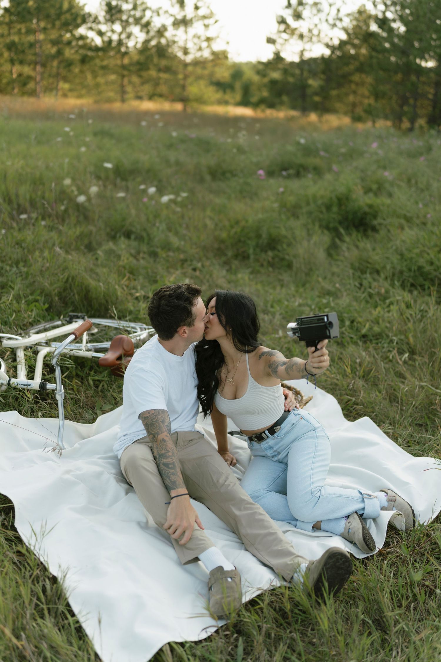 Couple sharing a kiss during a romantic outdoor picnic on a white blanket in a green meadow, taking a selfie with a camera.