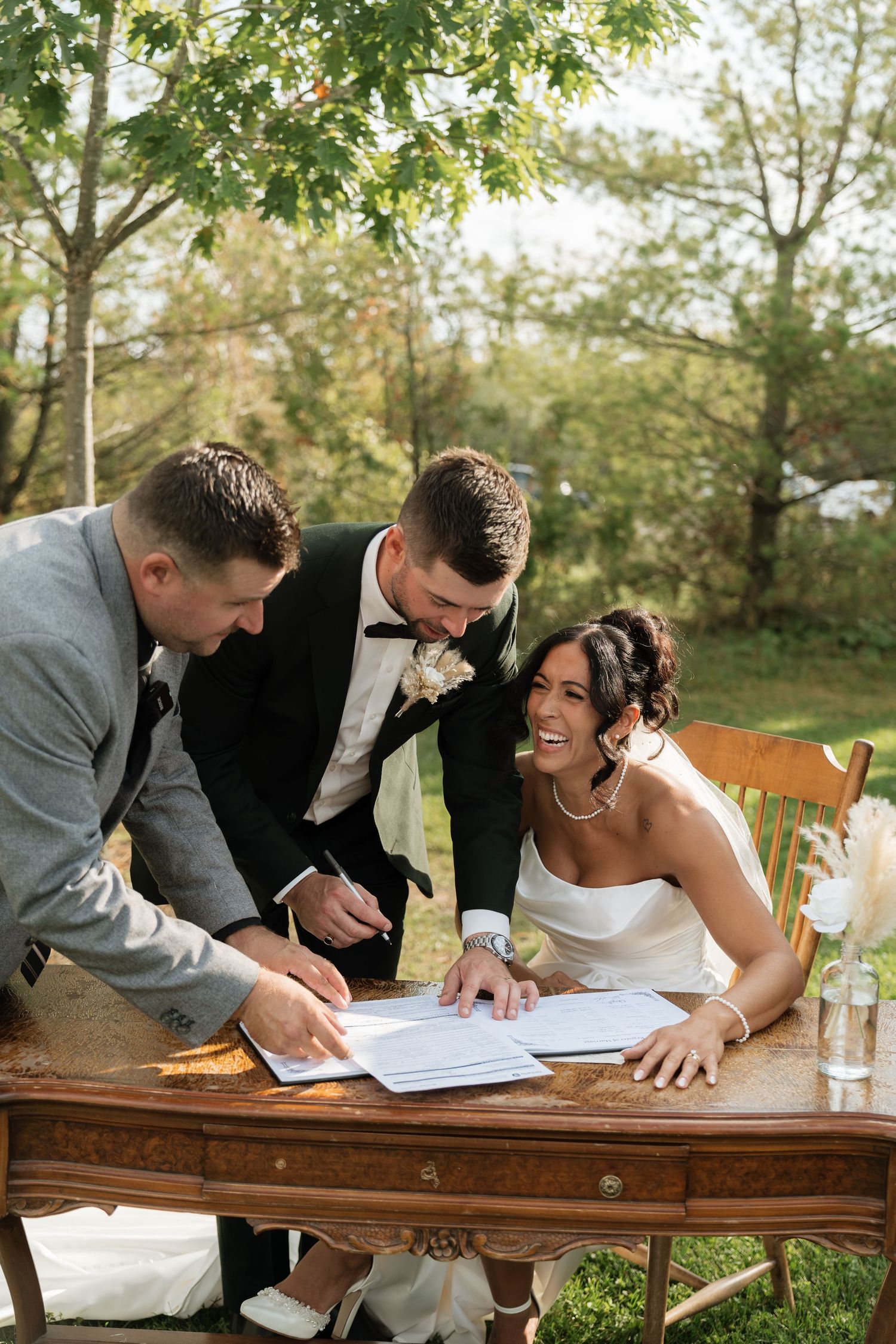 Wedding couple signing marriage license with witnesses at wooden table outdoors.