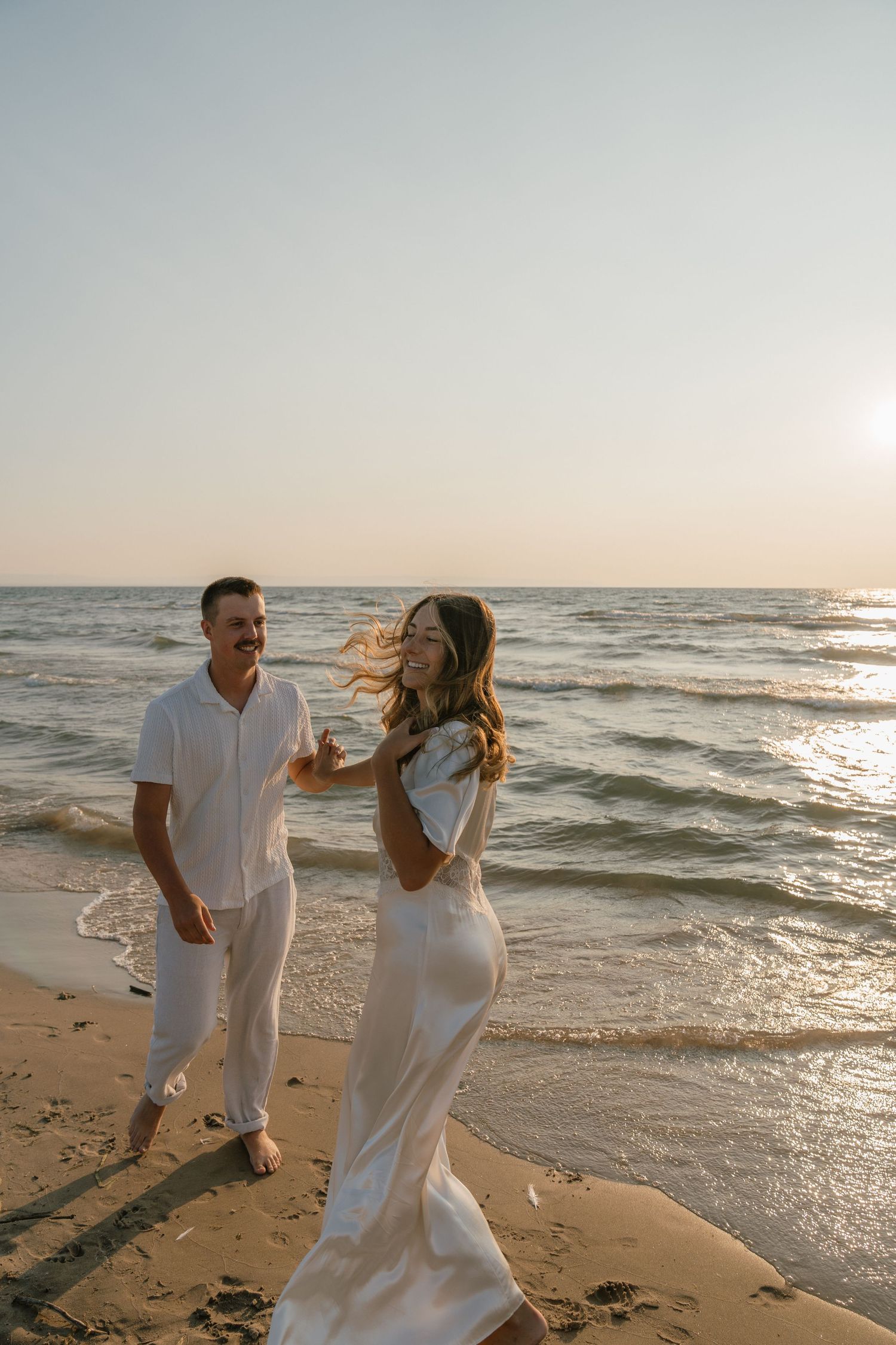 Couple in white outfits playing and running along beach shoreline at sunset.