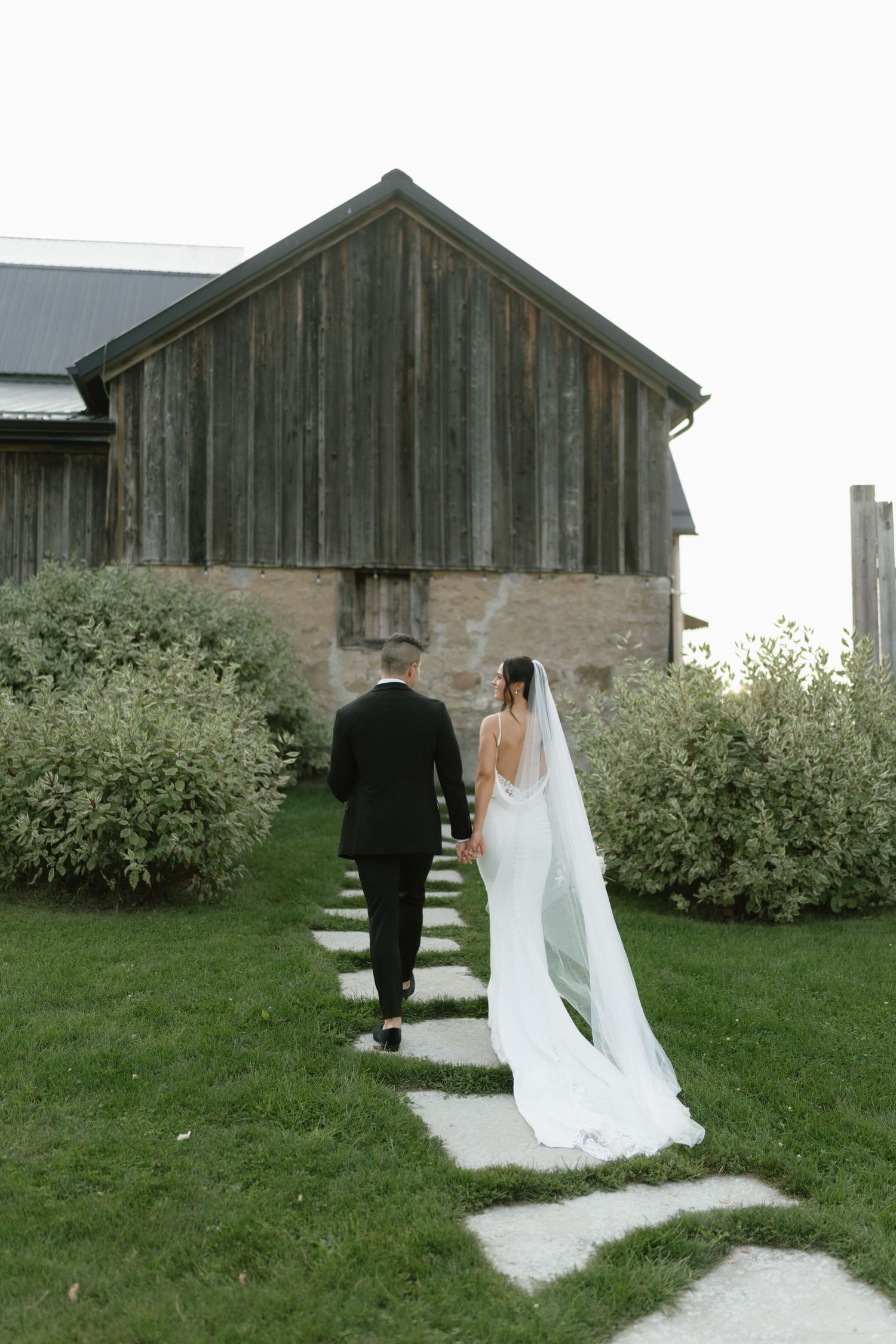 Bride and groom walking hand in hand toward a rustic wooden barn on their wedding day.