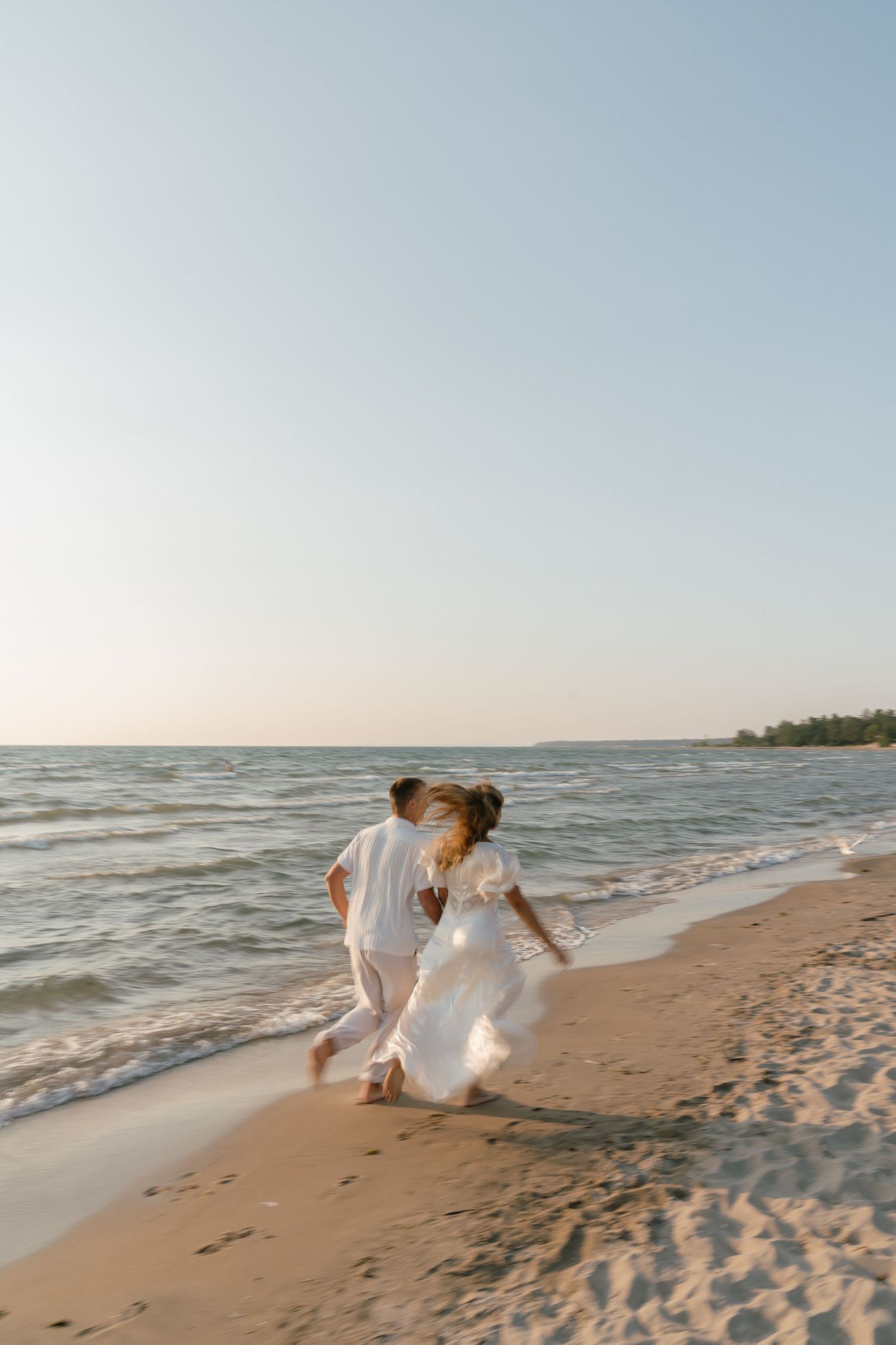 Couple walking along beach shoreline at sunset in casual wedding attire.