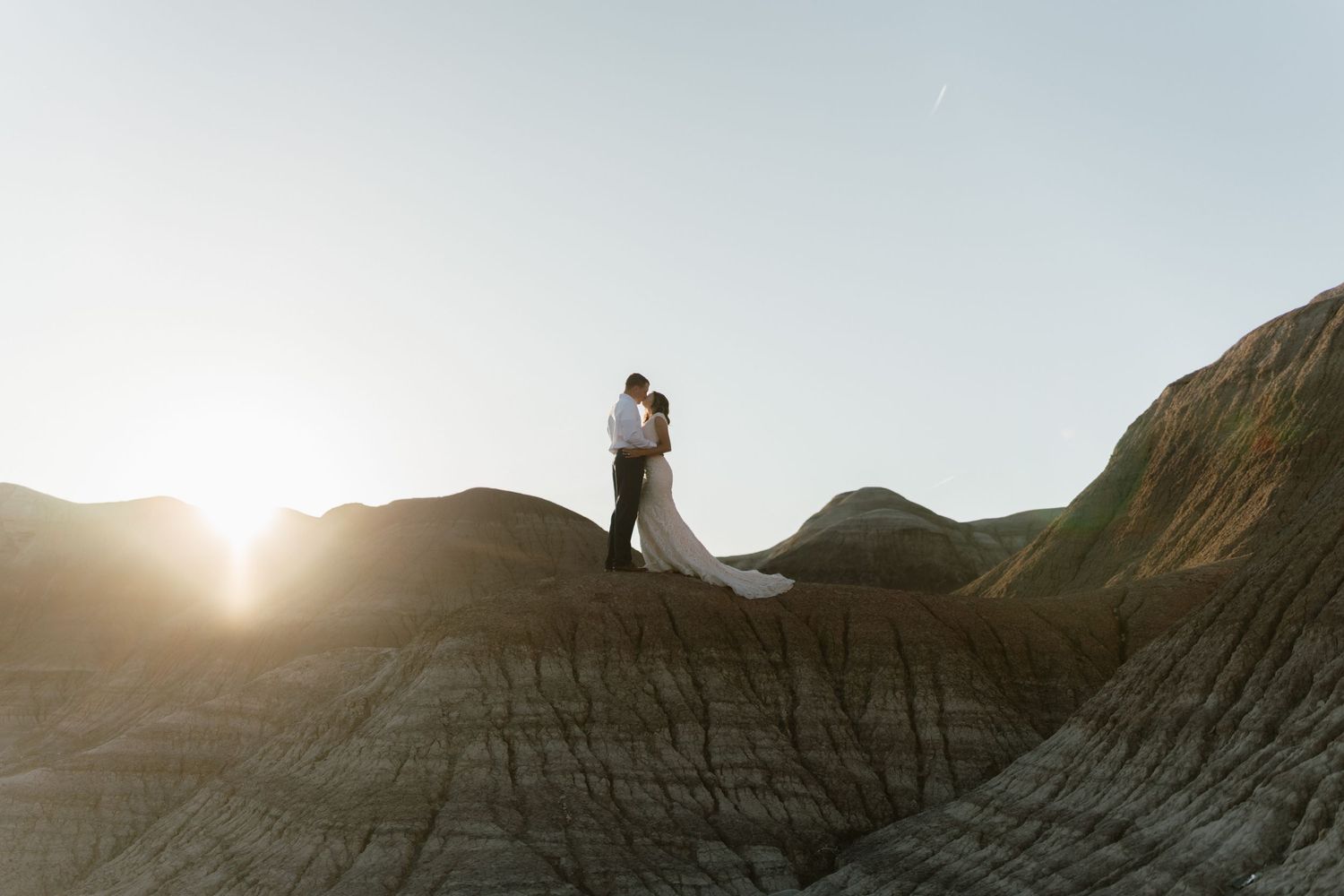 A couple stands silhouetted against a sunset on dramatic rocky terrain.