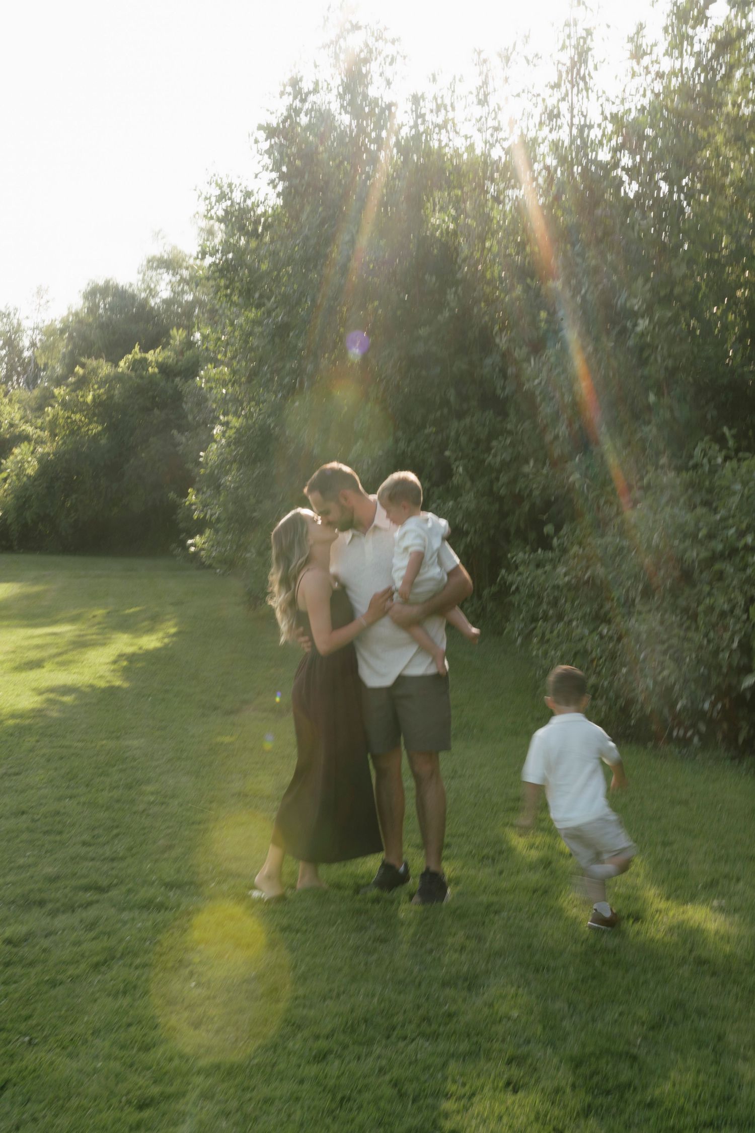 A family of four enjoys a sunny day on a green lawn, surrounded by lush trees with golden lens flare.