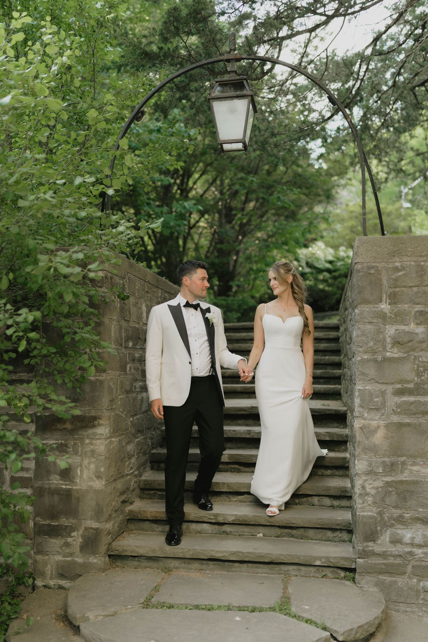 Couple descends stone steps under a vintage lamp post in elegant wedding attire.