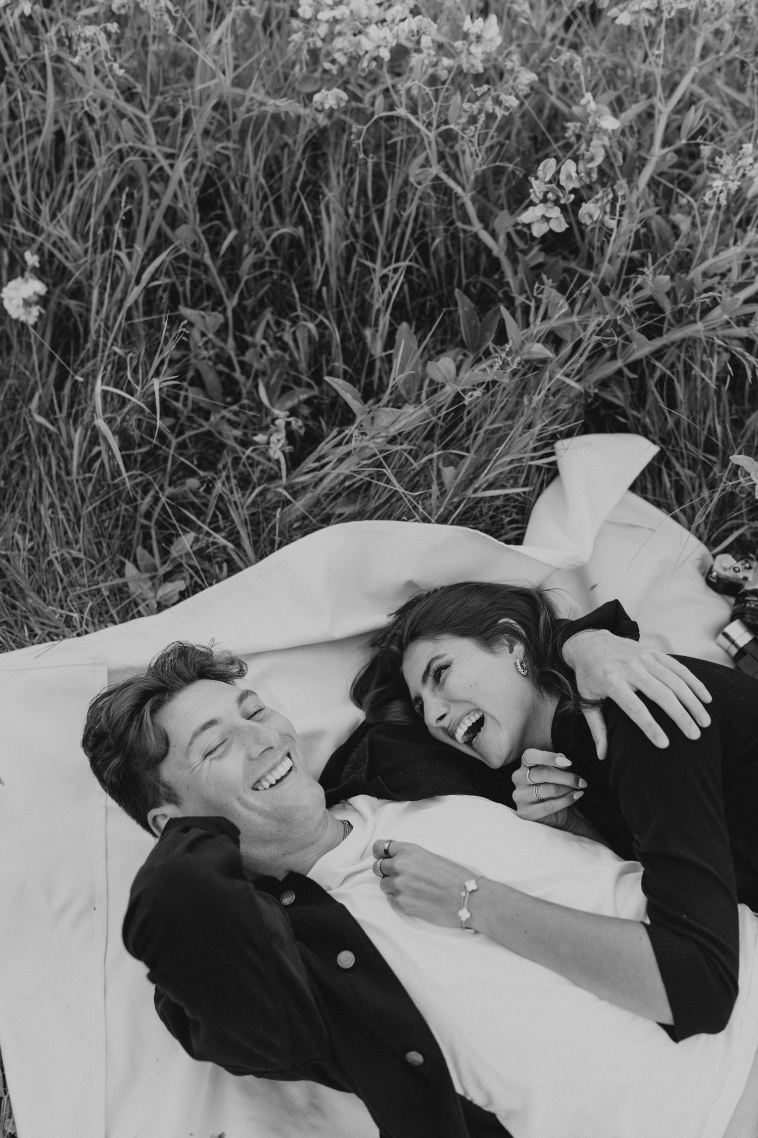 Black and white photo of a couple lying on white sheets in a field of flowers.