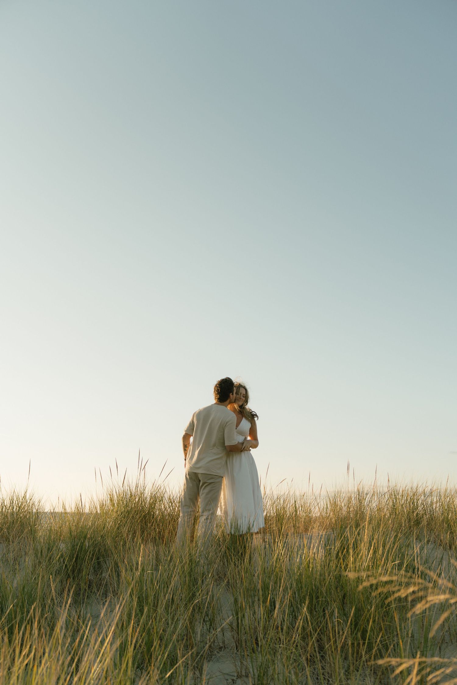 A couple walks through tall grass against a sunset sky in an intimate outdoor moment.