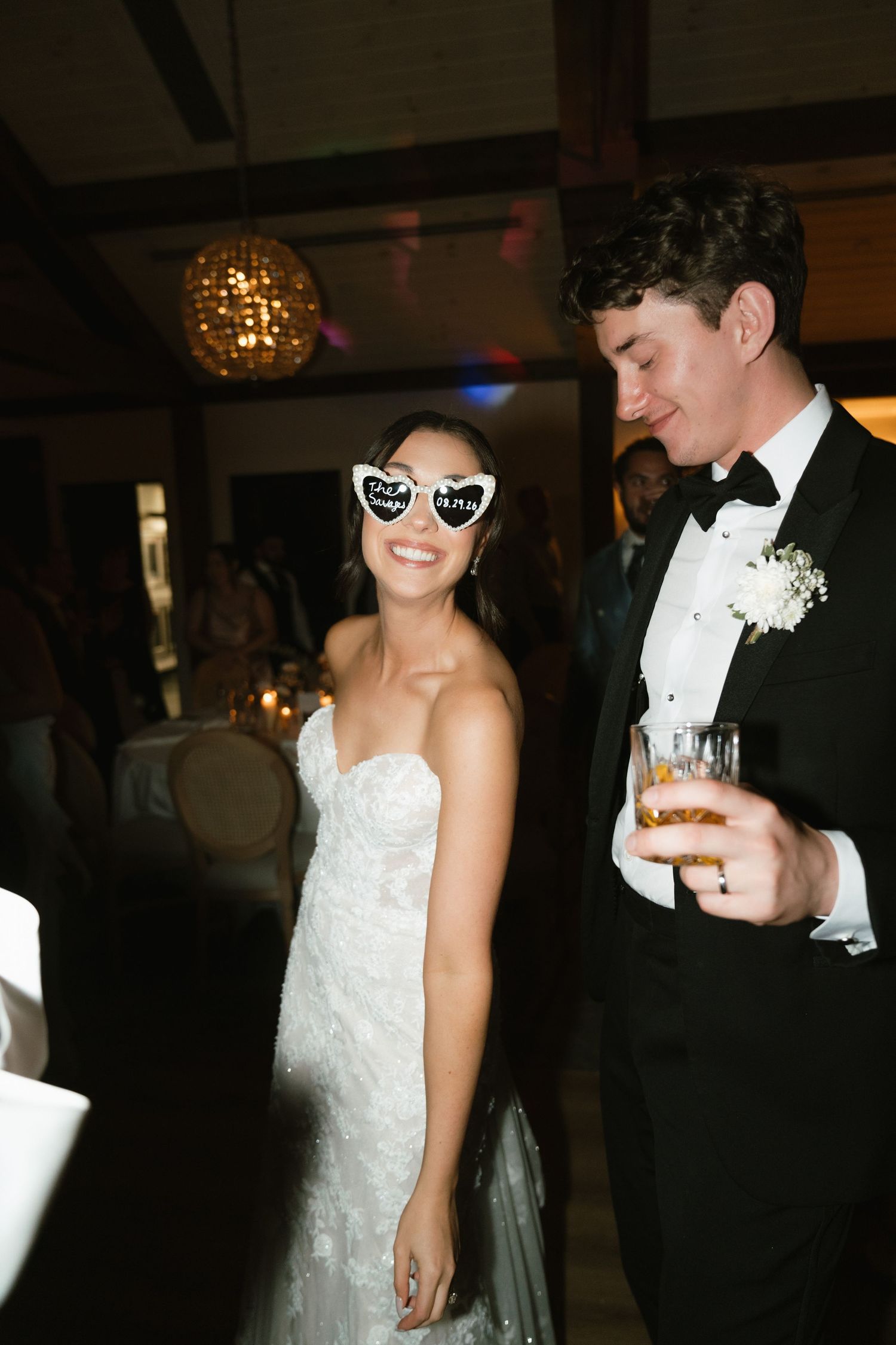 Bride wearing heart-shaped bridal sunglasses laughs with groom in tuxedo holding drink at wedding reception.