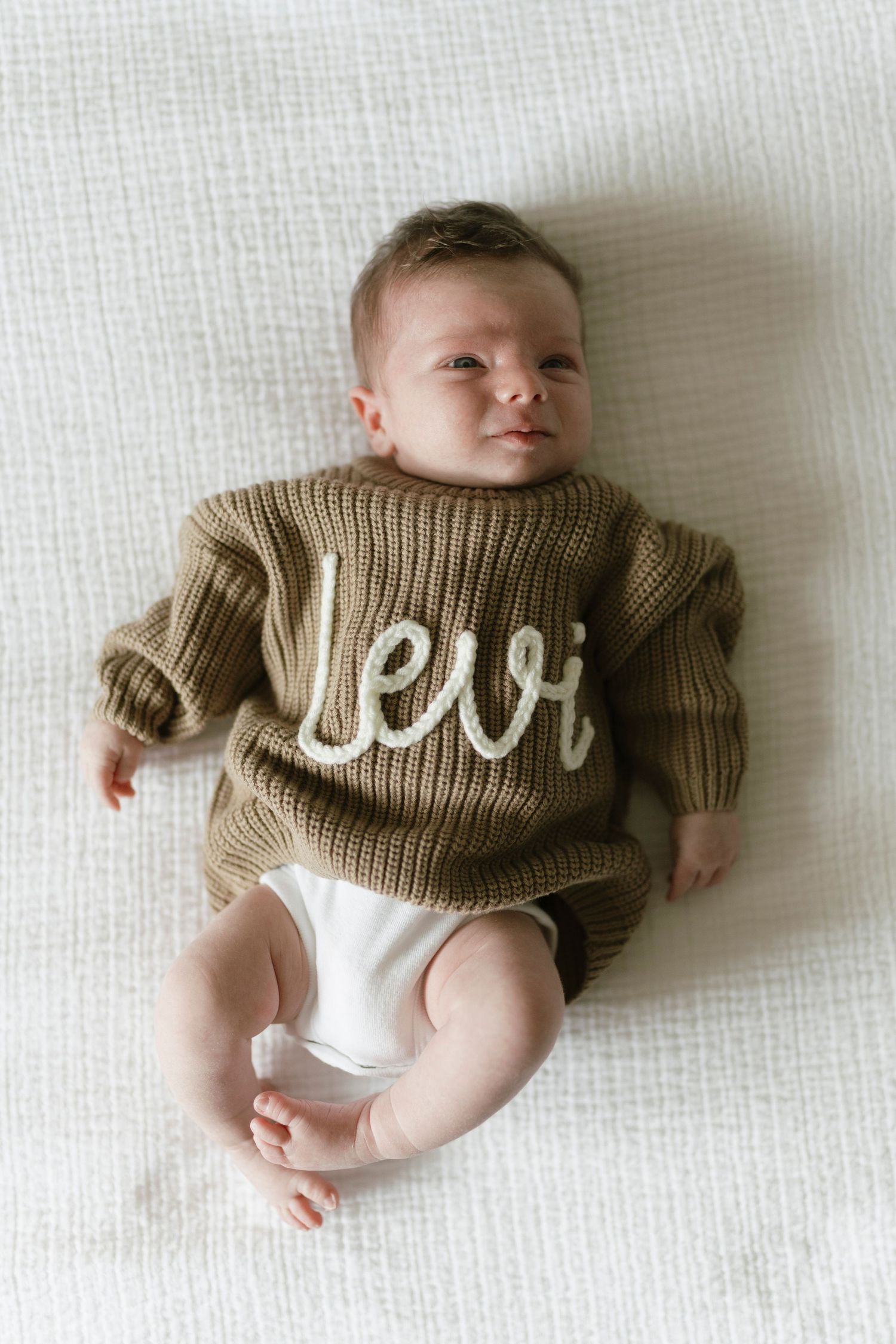 Baby wearing an olive green knit sweater with the name Levi embroidered, lying on a white blanket.