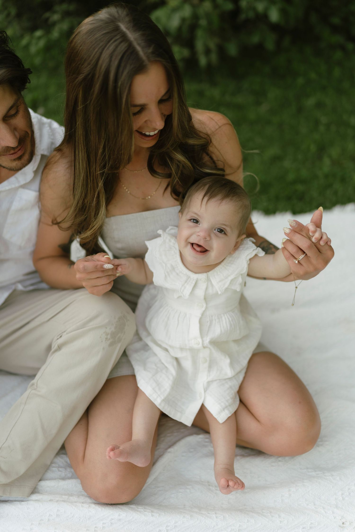 Happy baby in white dress laughs while parents hold hands outdoors on white blanket in lush green garden.