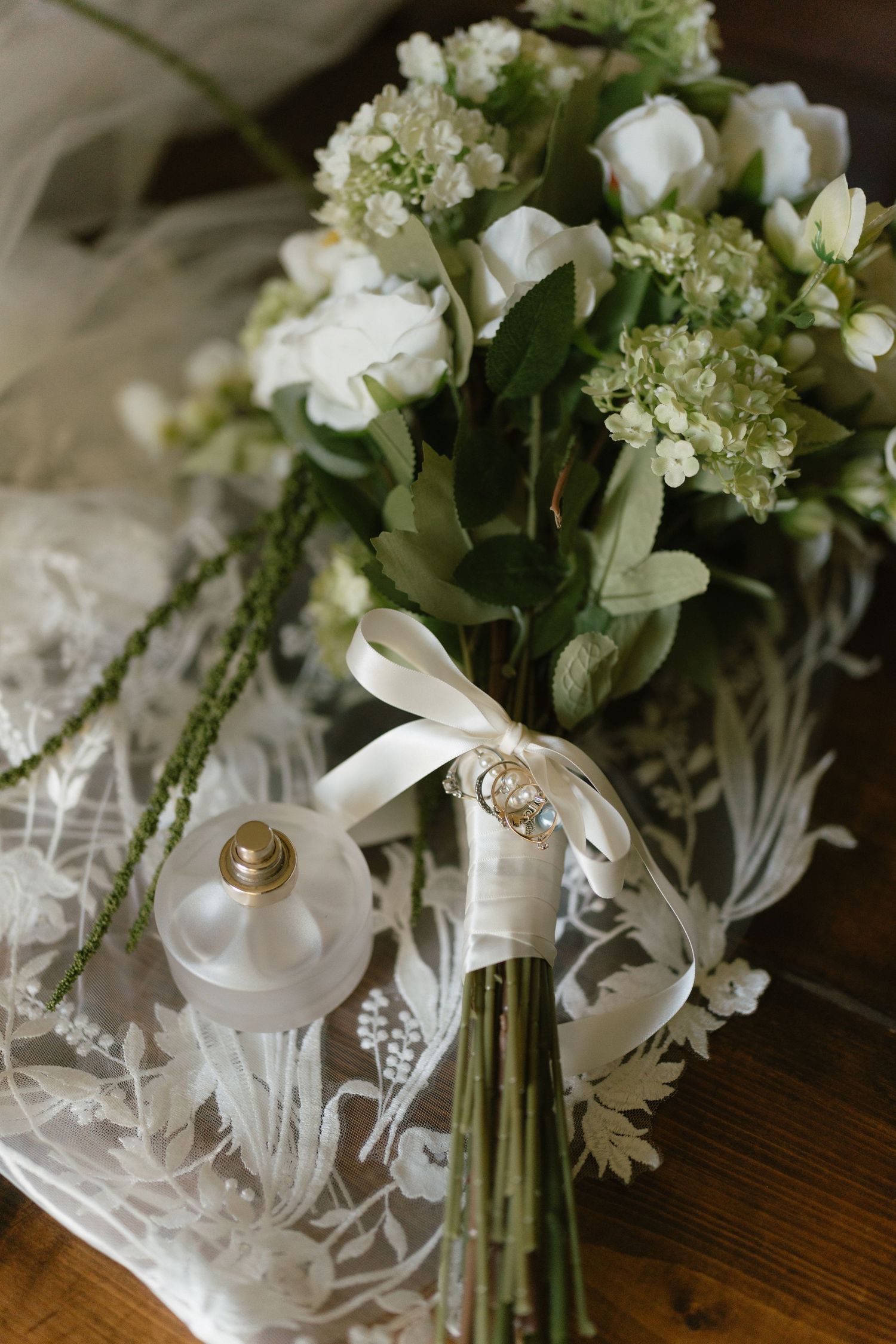 White floral bouquet with ribbon and wedding rings displayed on lace fabric.