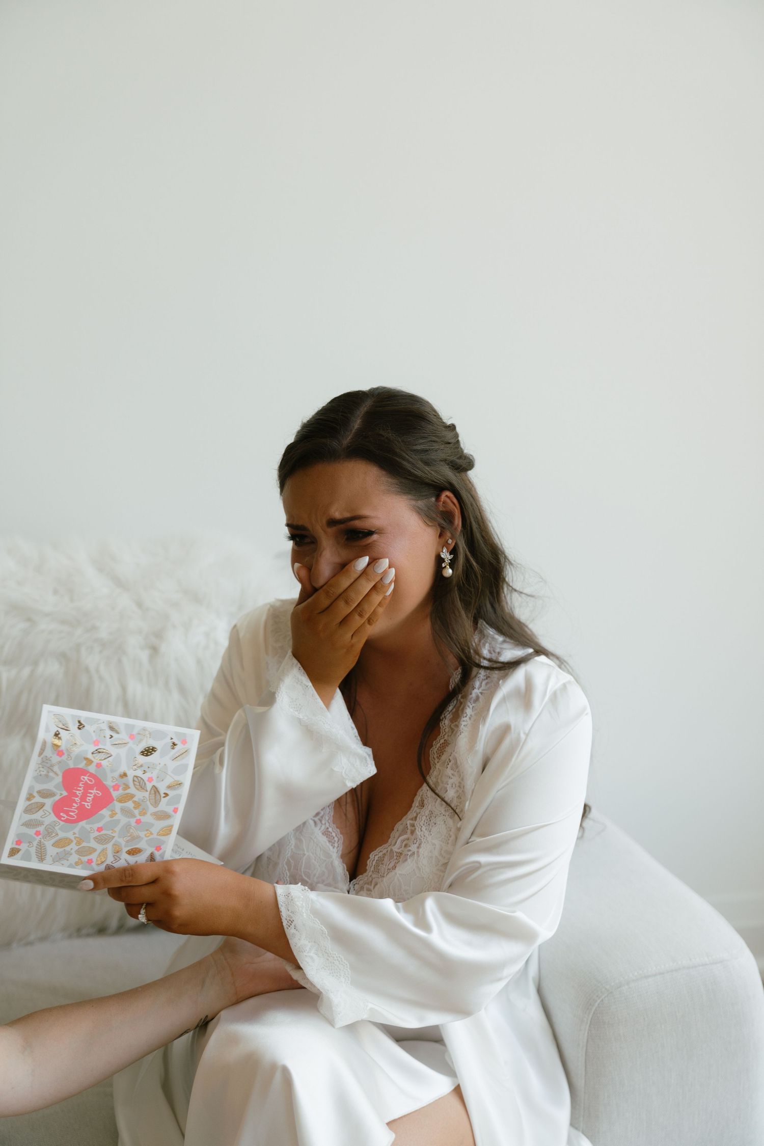 Someone in a white robe reacts emotionally while reading a card on a white couch.