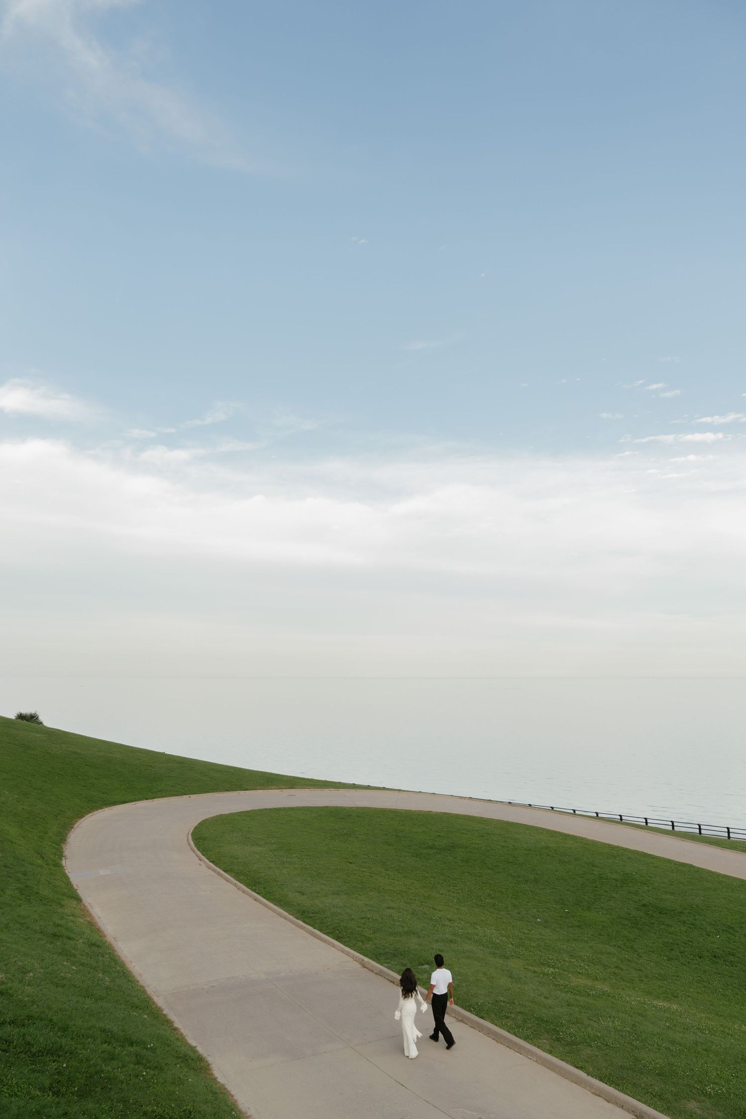Couple walking hand in hand along a curved path overlooking a serene ocean horizon under a soft blue sky.