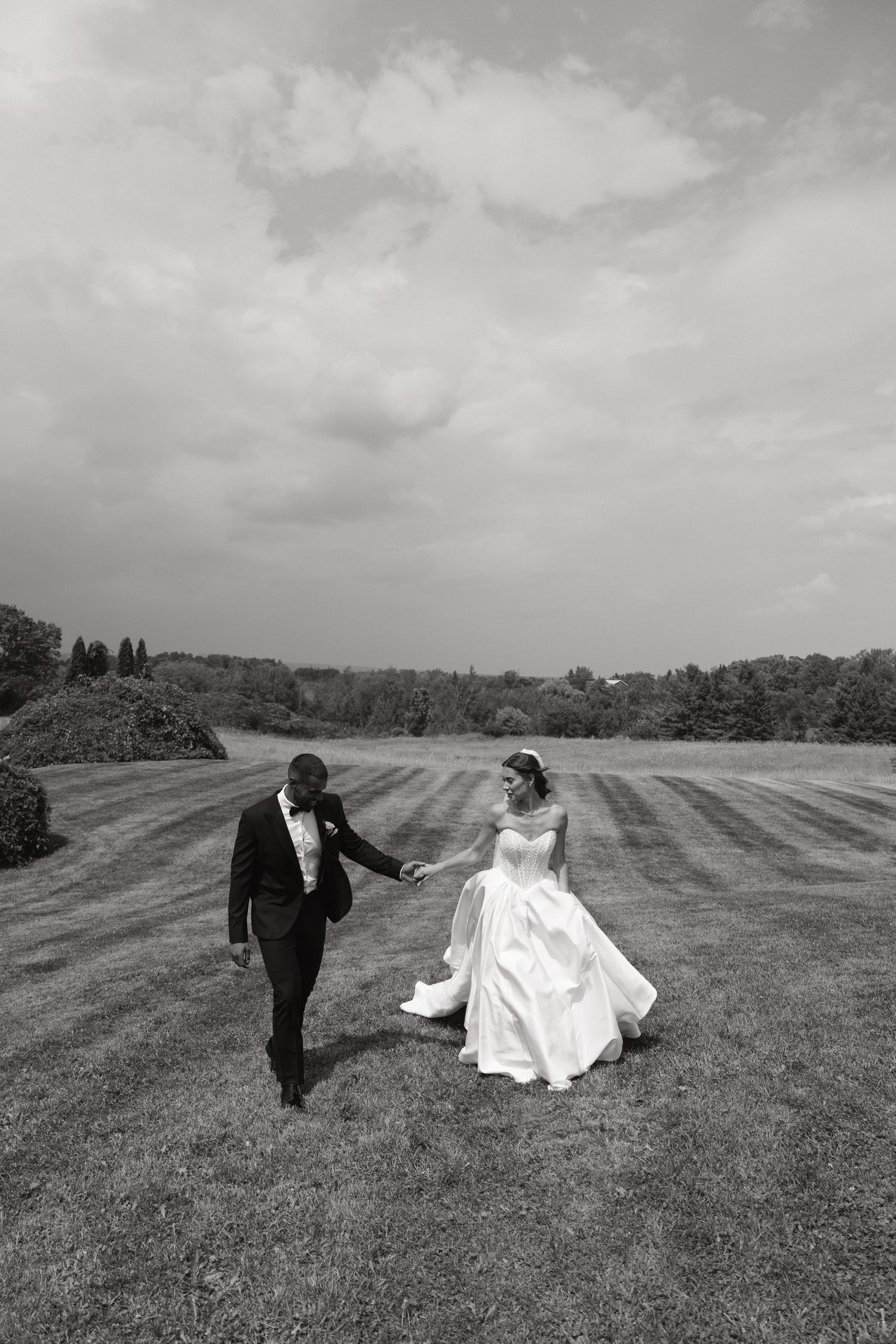 Black and white photo of couple running across striped lawn under cloudy sky.