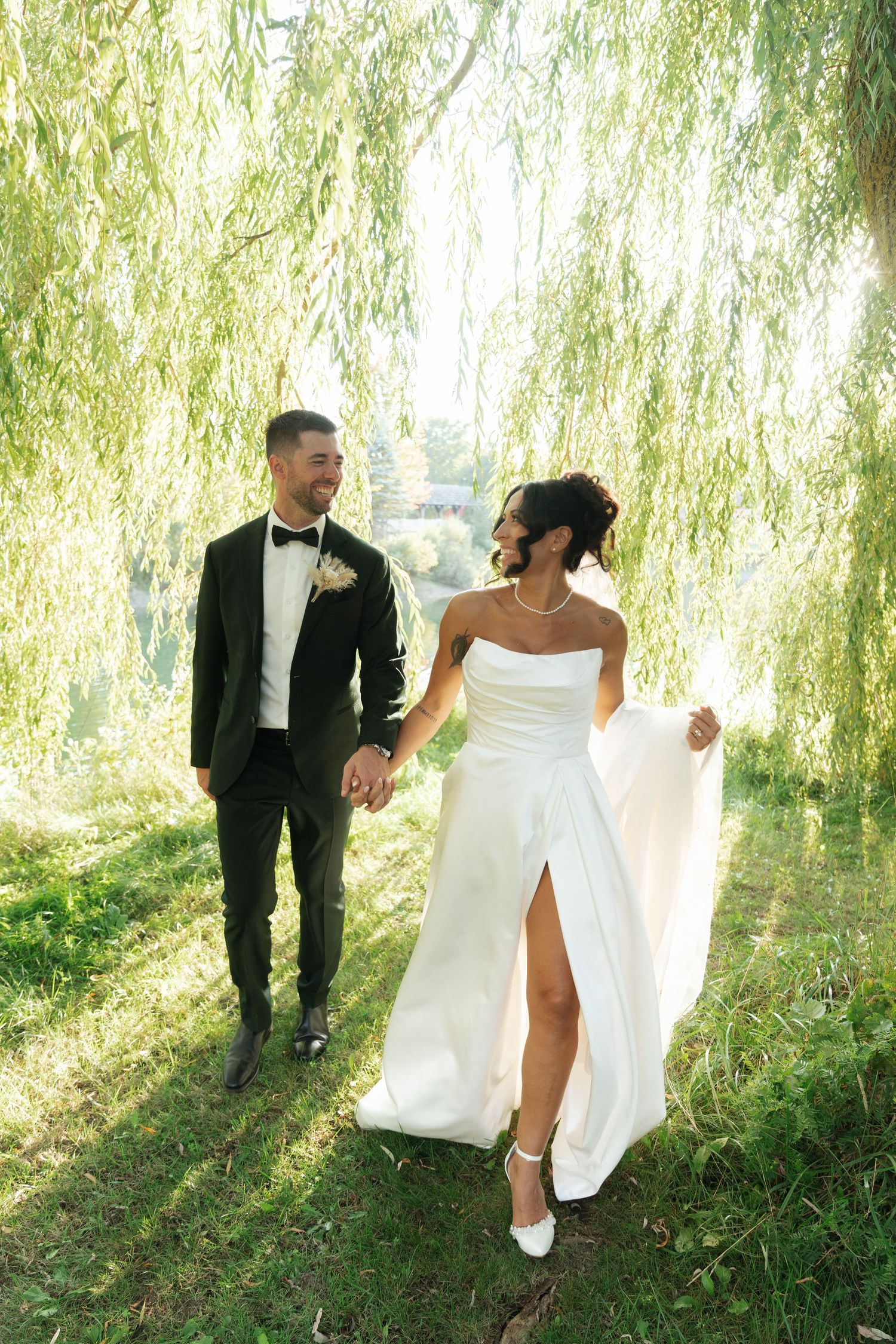 Newlyweds walk beneath a romantic weeping willow tree, with the bride in an elegant white dress with a high slit.