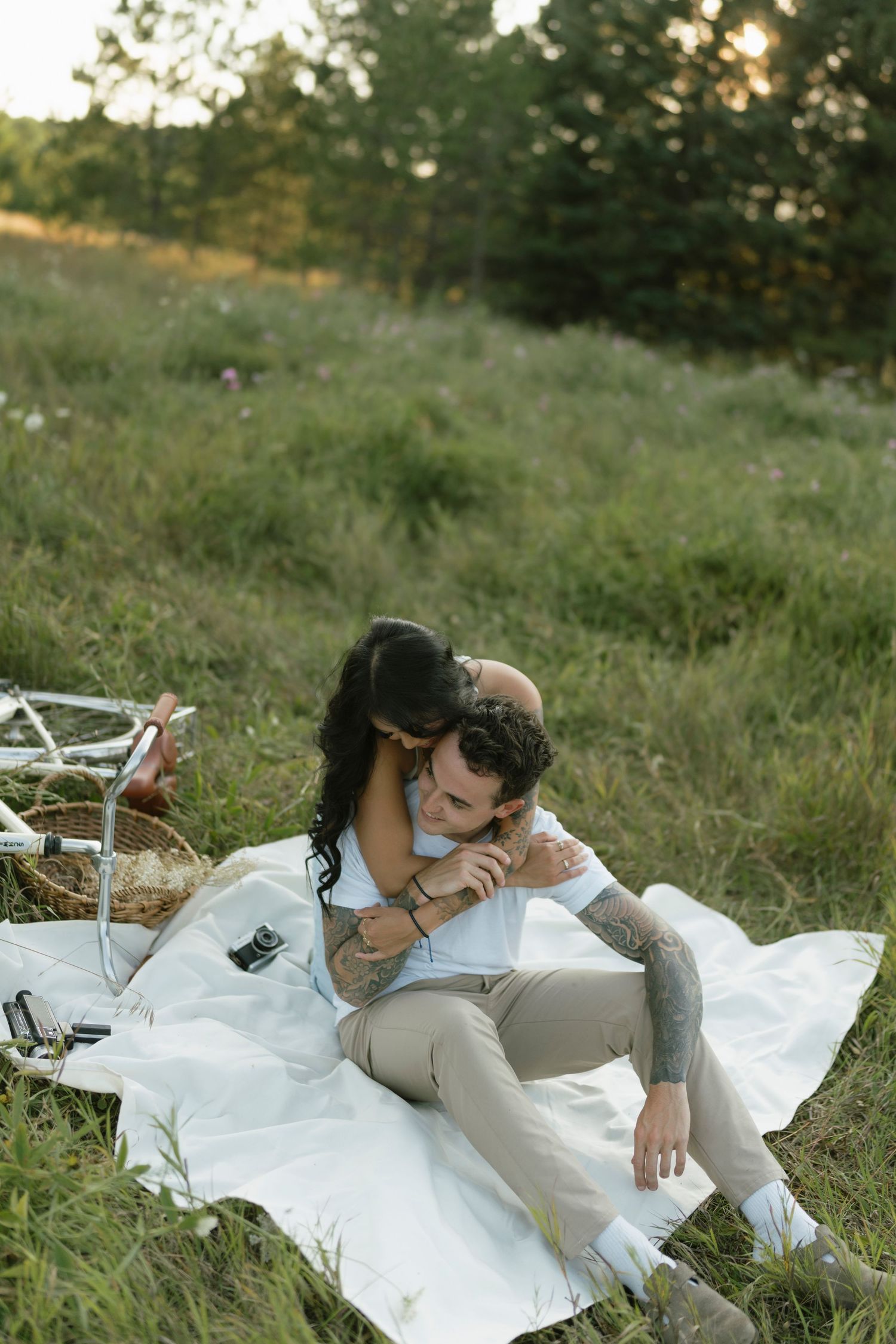 Couple sharing a romantic picnic on a white blanket in a green meadow, with a bicycle and basket nearby at sunset.