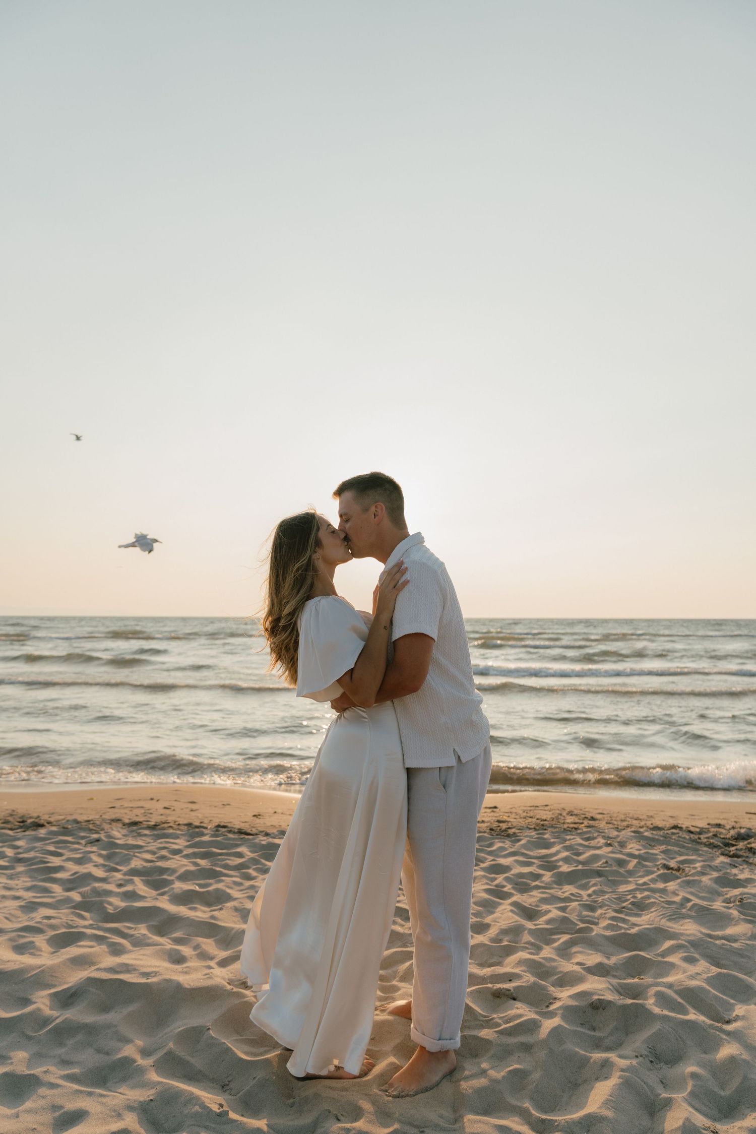 Romantic beach photoshoot with couple in white outfits standing on sandy shore at dusk.